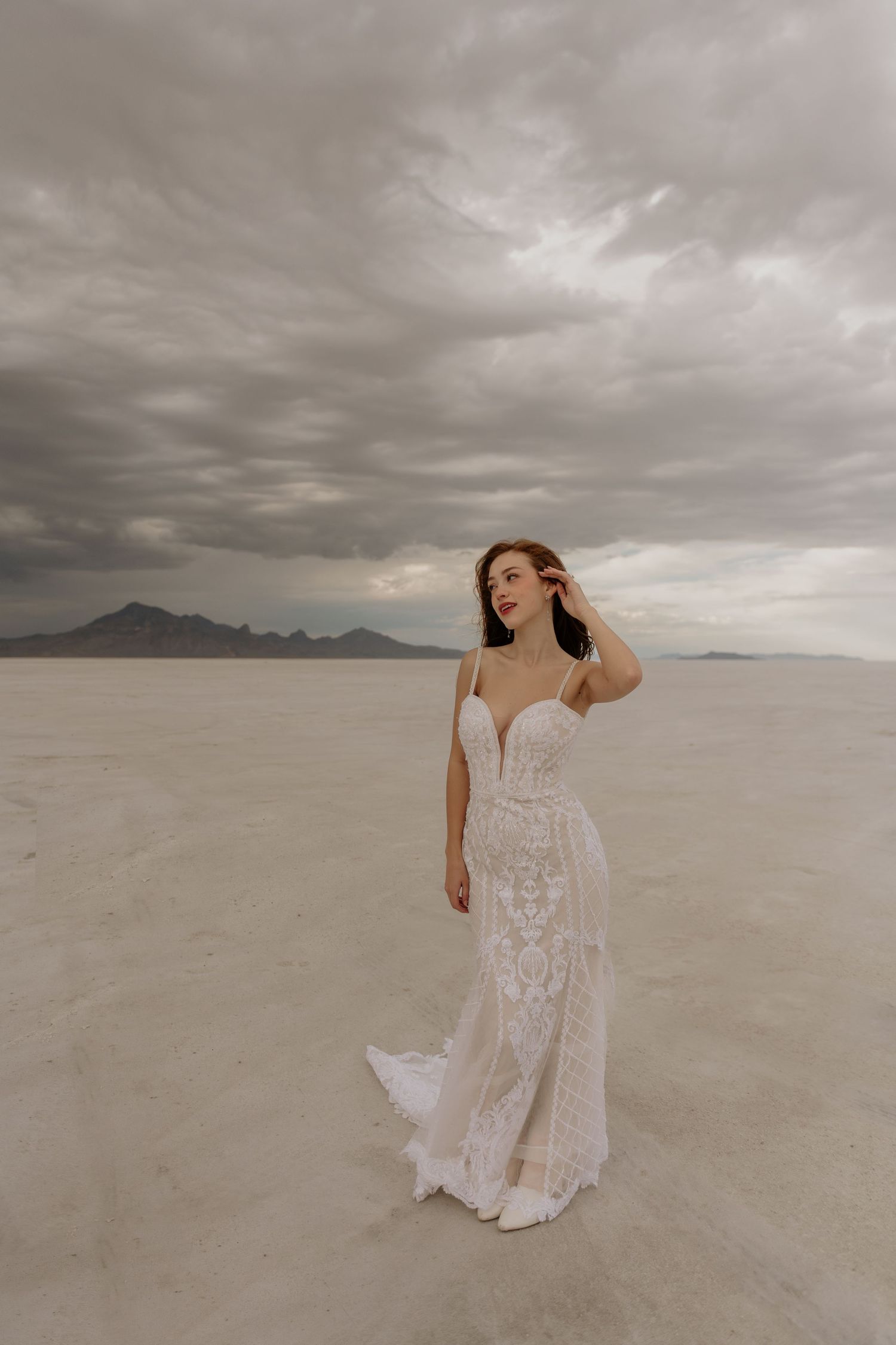 A bride in a white lace wedding dress stands on the Bonneville Salt Flats under dramatic stormy clouds.