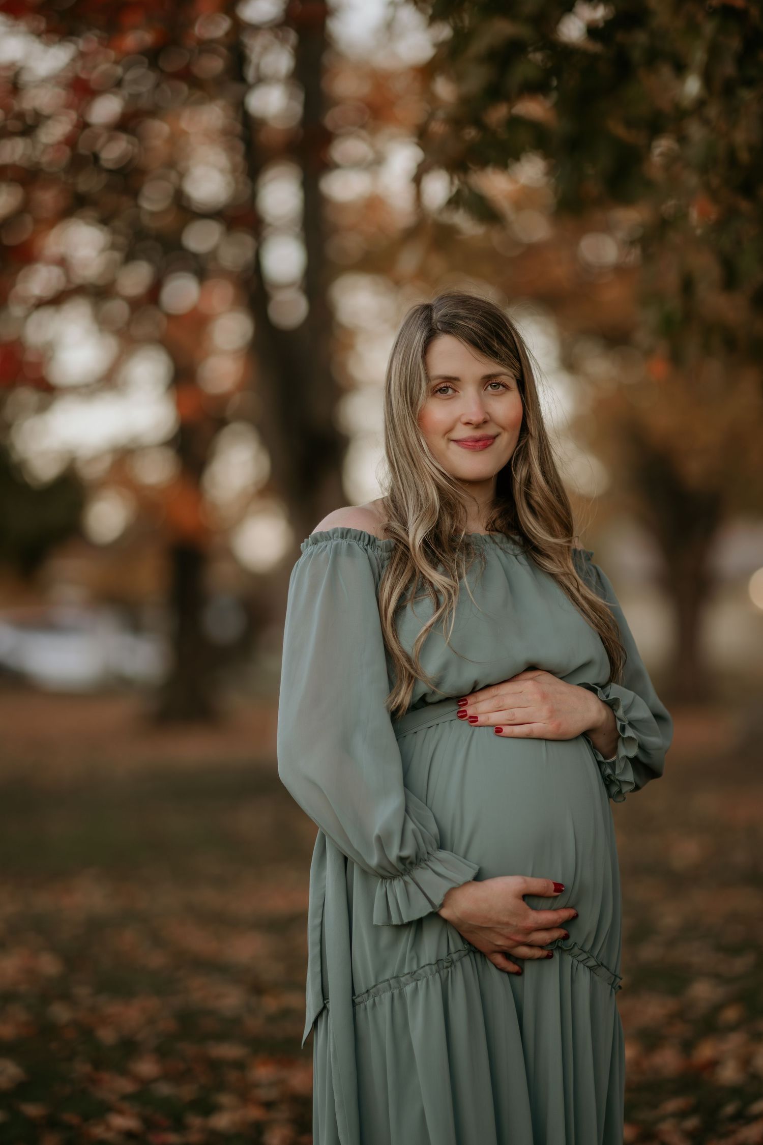 Fall maternity portrait session featuring woman in flowing sage green dress among autumn leaves.