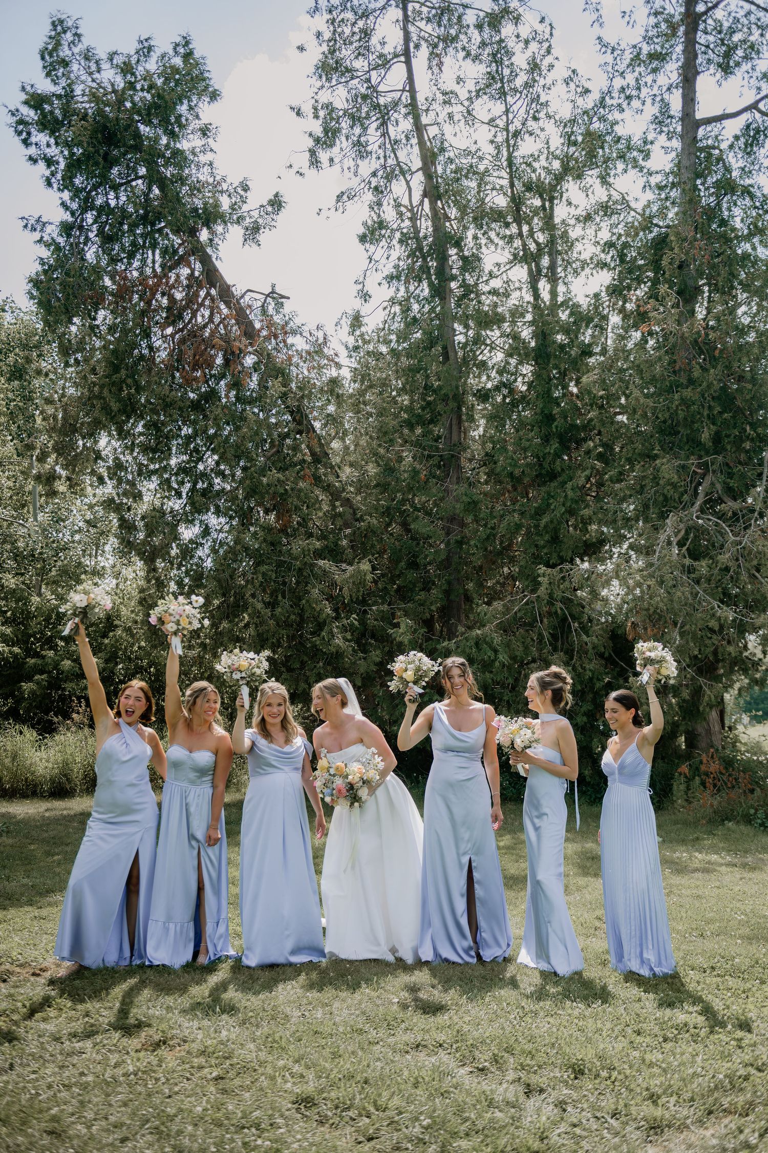A wedding party in pale blue dresses posing together on a grassy field with trees in the background.