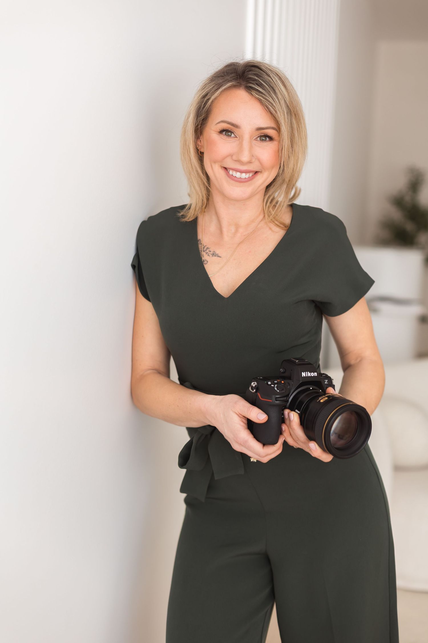 Professional photographer in dark green outfit poses with camera in bright white studio setting.