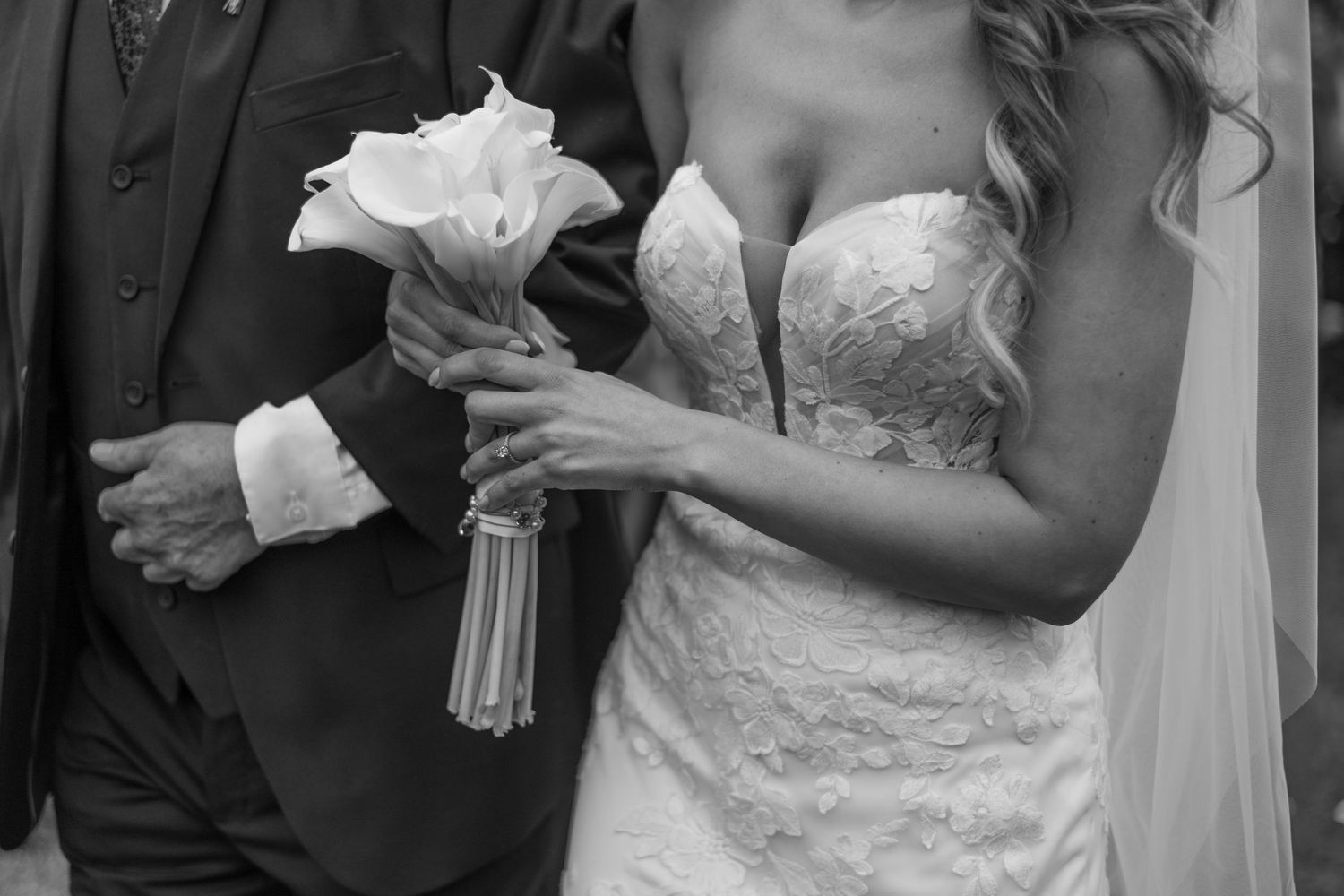 Black and white photo of bride in lace gown holding calla lily bouquet with groom in suit at wedding ceremony.