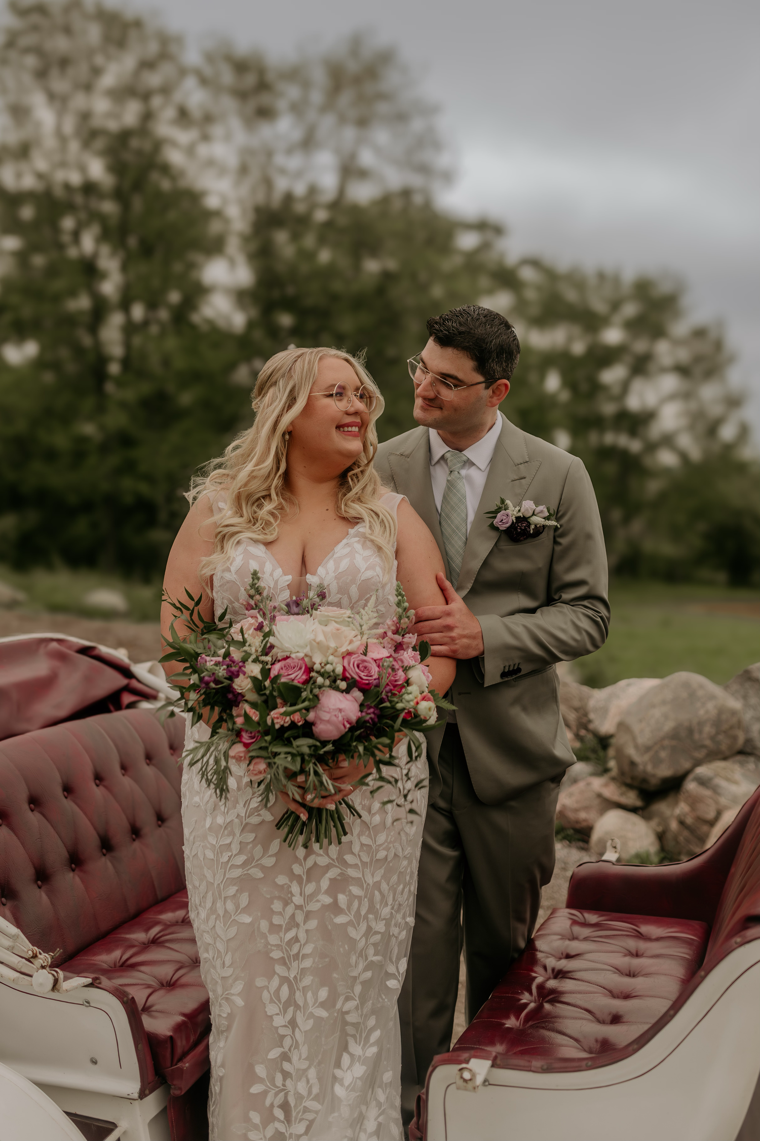 Wedding couple sitting on vintage burgundy sofa outdoors with floral bouquet.