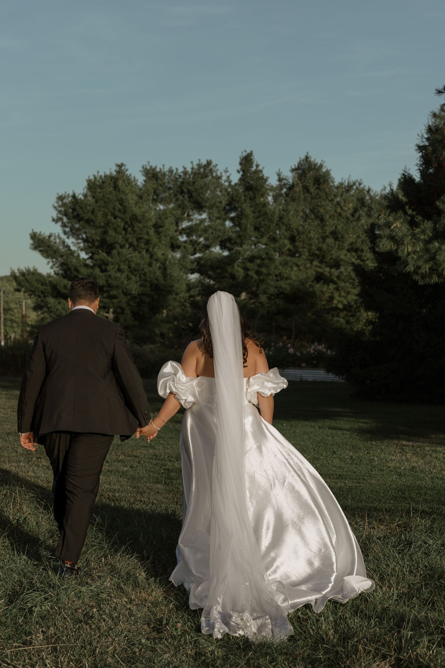 Bride and groom walk through a grassy field with evergreen trees under blue sky during wedding photoshoot.