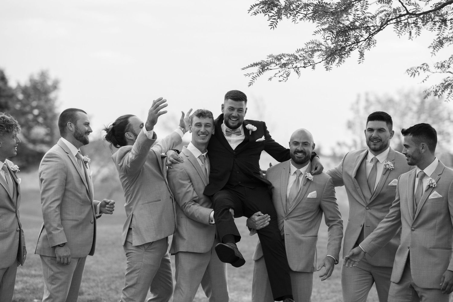 Groomsmen in gray suits celebrate joyfully outdoors during wedding festivities.
