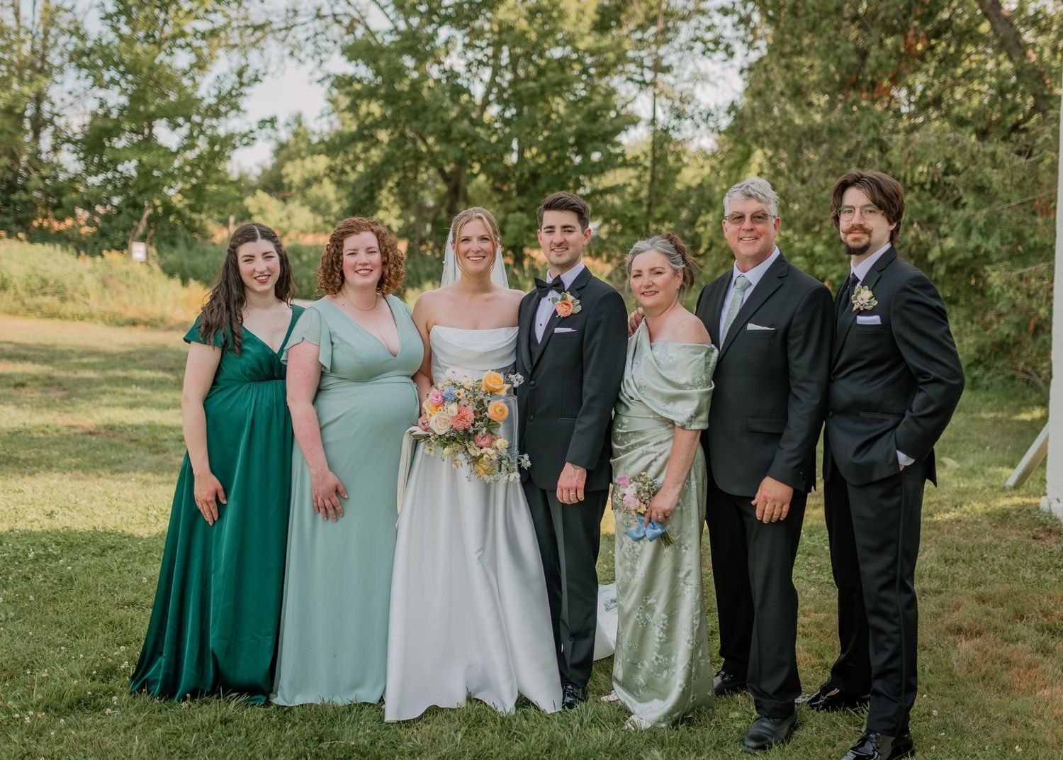Wedding party in formal attire poses together outdoors with bridesmaids in blue and green dresses.