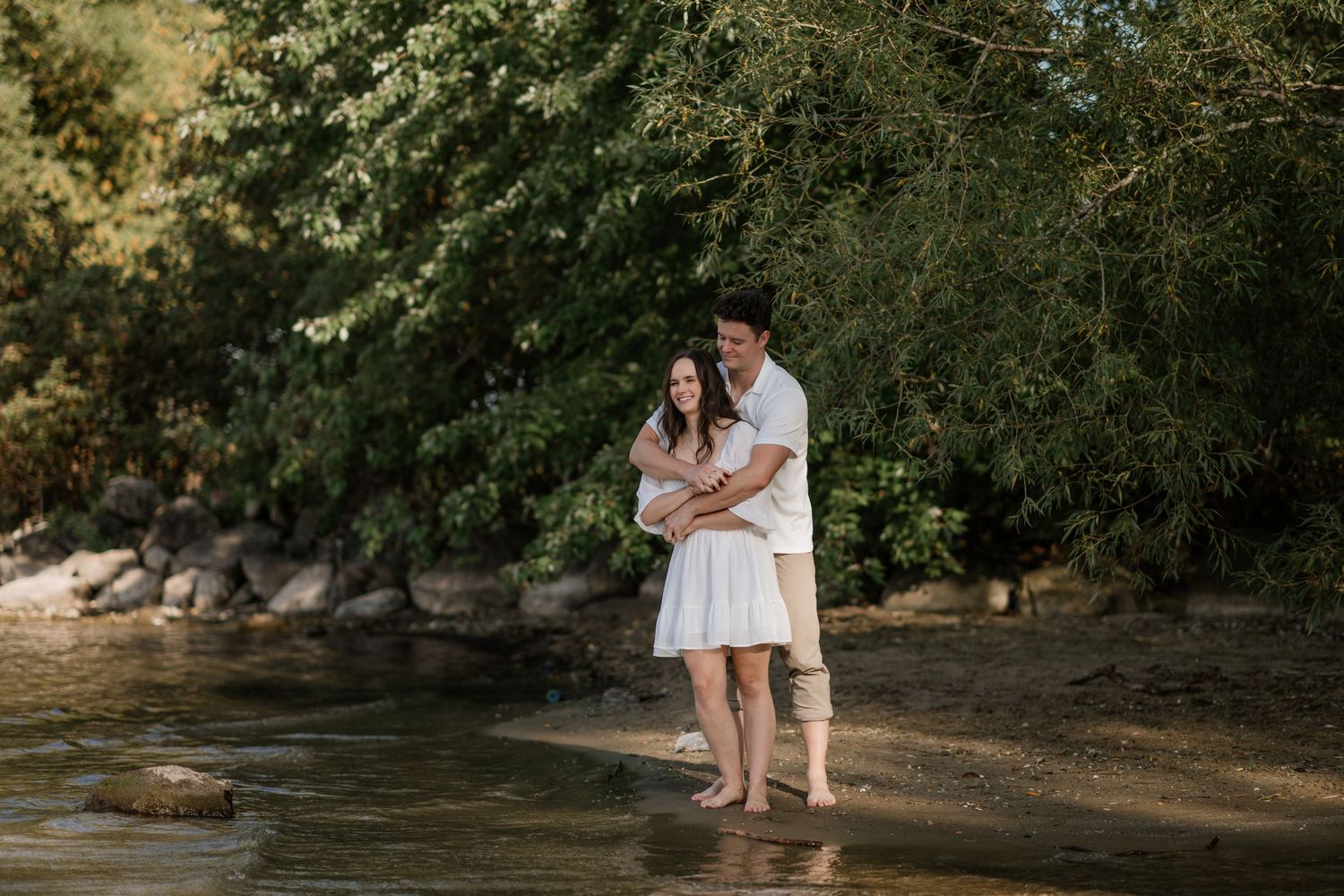 A couple embraces while standing in shallow water near a rocky shoreline.