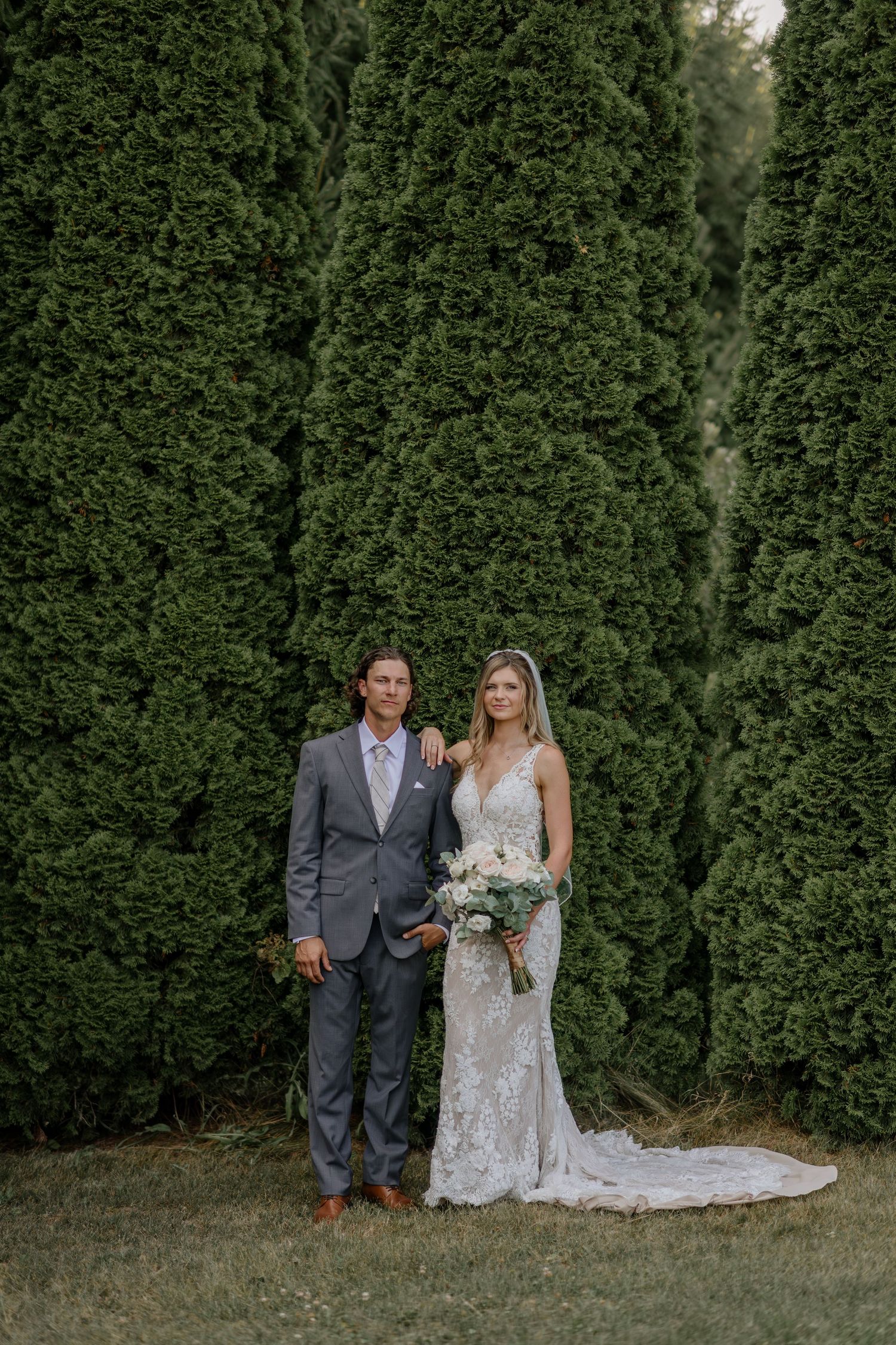 Wedding couple posing against a backdrop of tall green cypress trees.