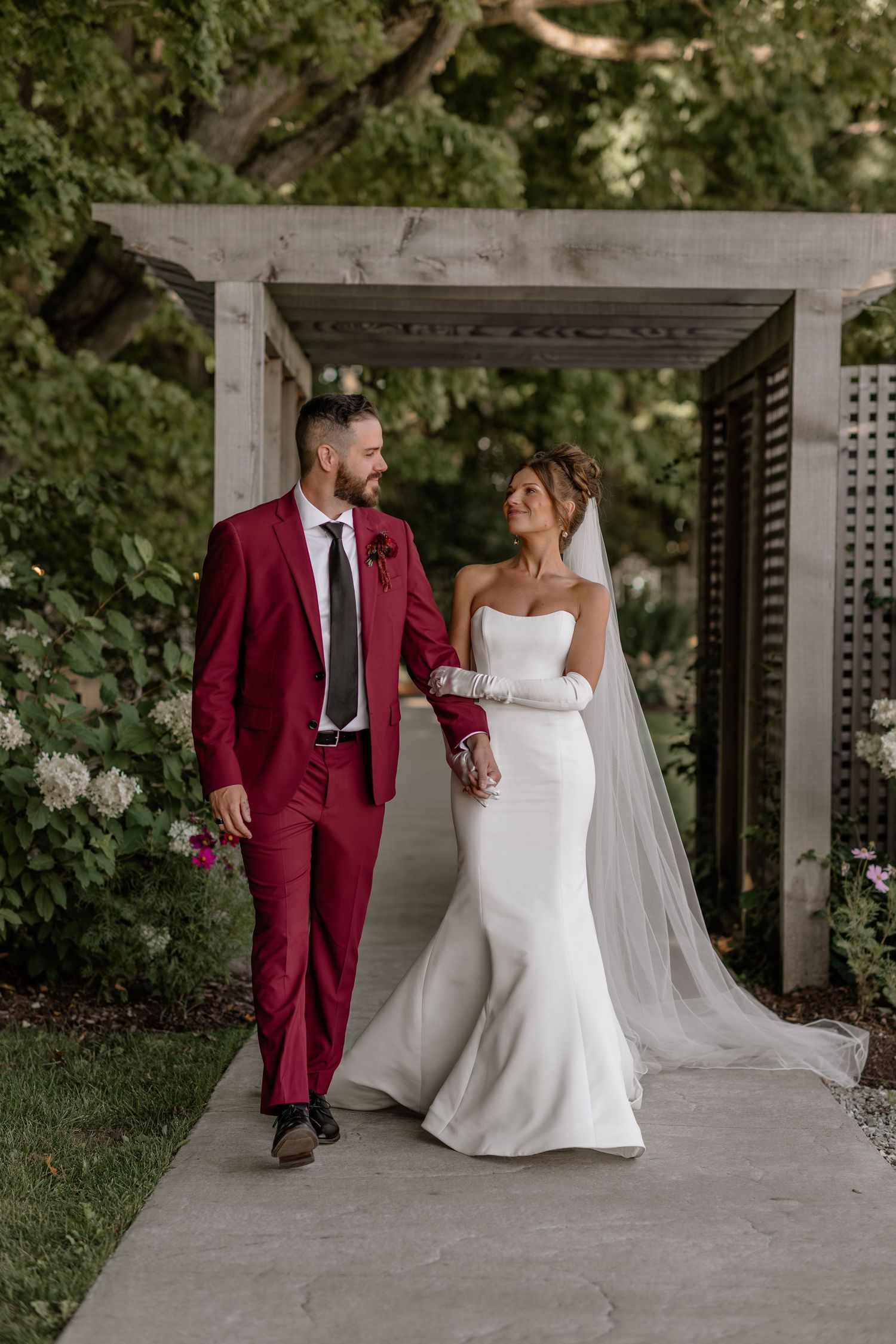 A couple in a burgundy suit and white wedding dress pose together under a pergola.