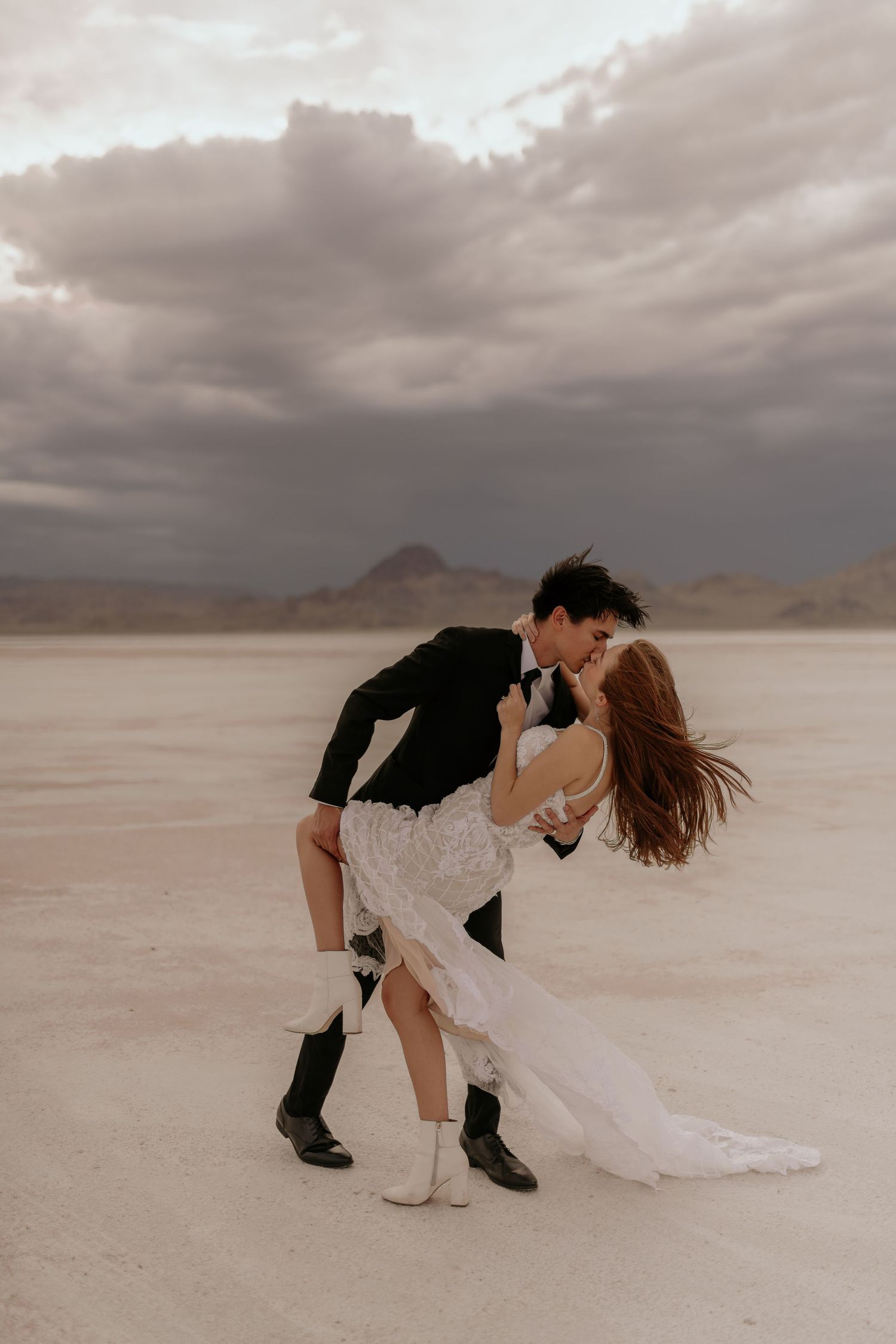 A romantic couple dips and embraces on a dramatic salt flat landscape under moody storm clouds.