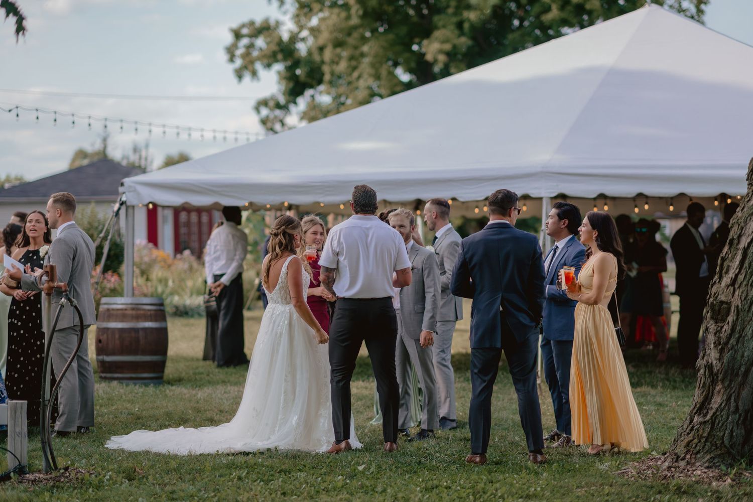 Wedding guests gather under white tents at an outdoor reception venue during sunset.