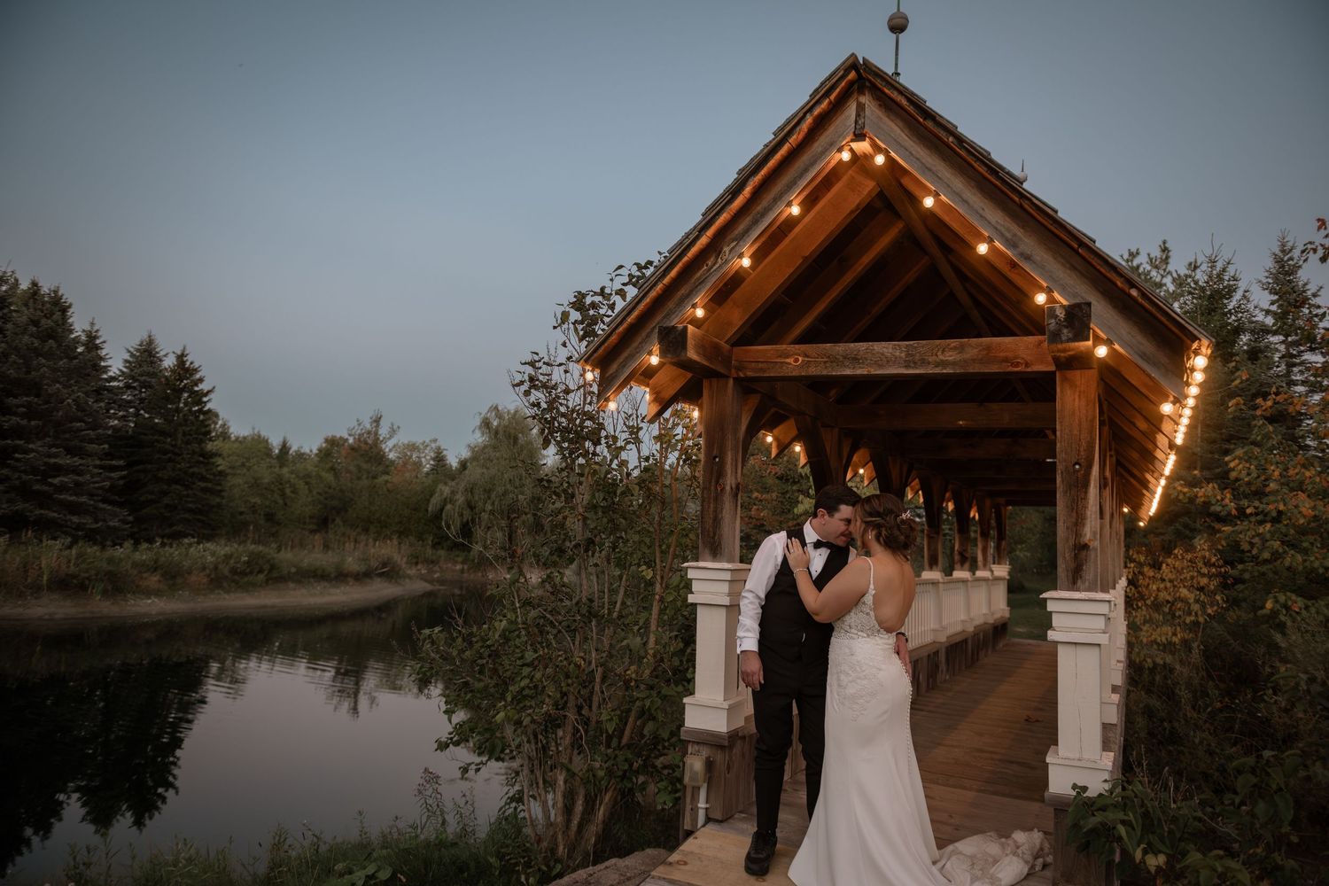 A romantic outdoor wedding ceremony at a rustic wooden pavilion with string lights and natural lake backdrop.