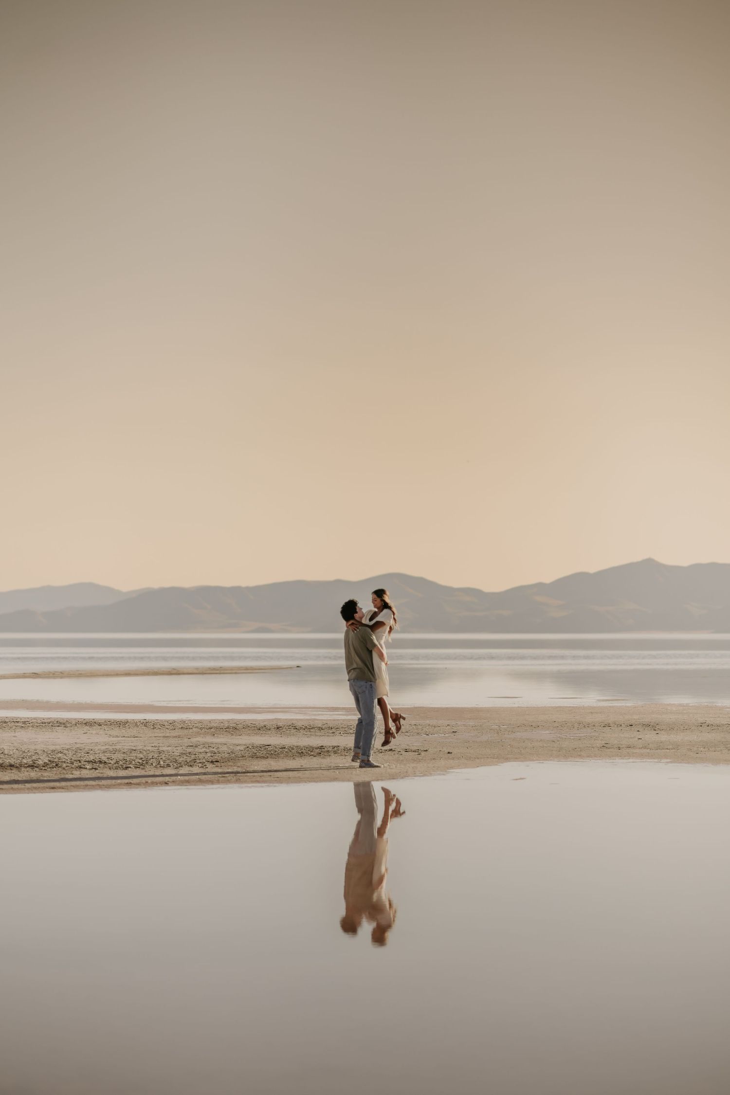 A couple dances together on a serene salt flat with mountains in the distance during sunset.