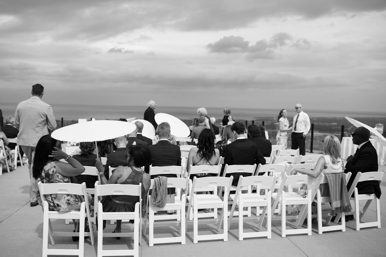 Black and white photo of guests seated in white chairs at an outdoor wedding ceremony with umbrellas and ocean view.