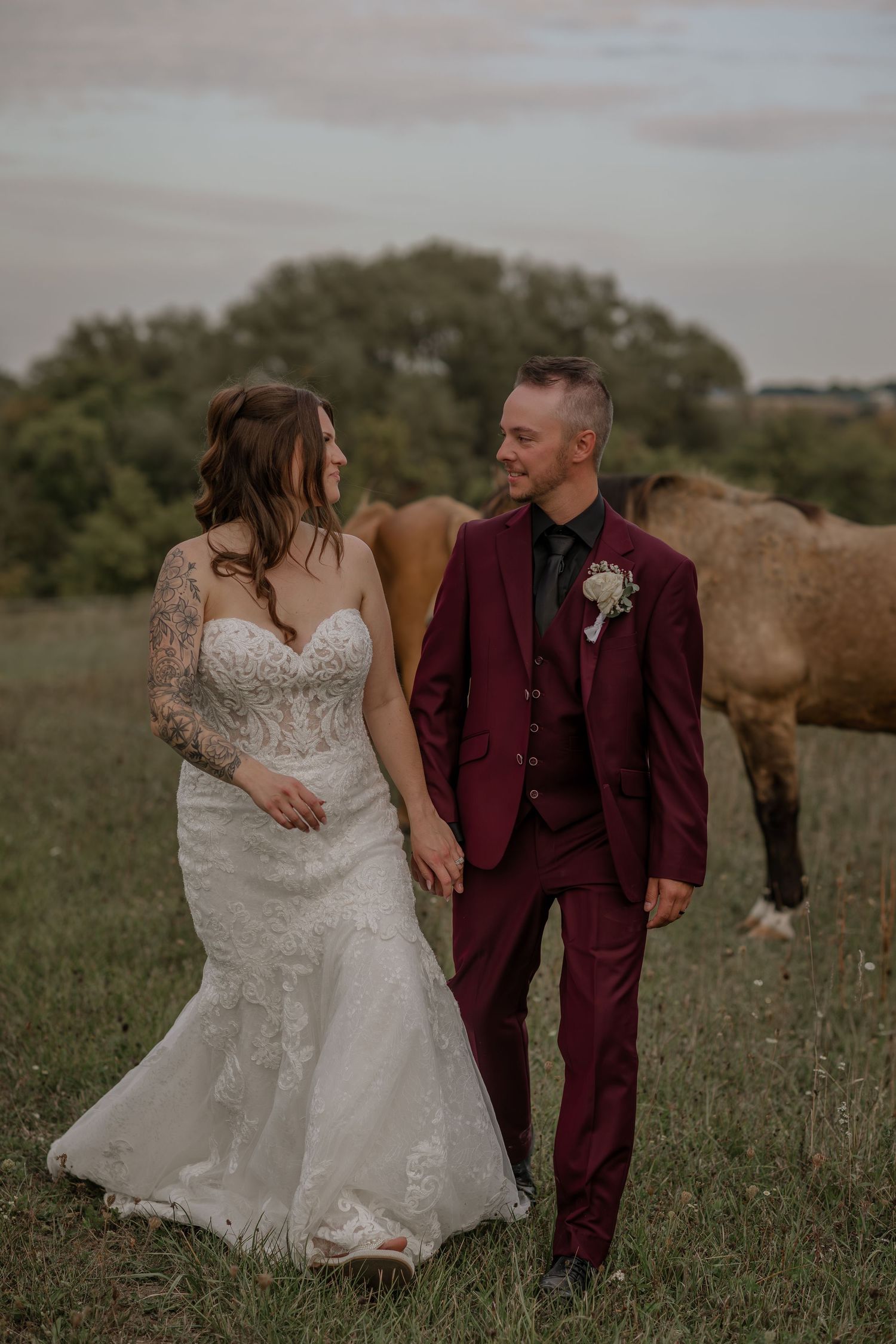 Couple in burgundy suit and white dress share intimate moments in a rustic field setting.