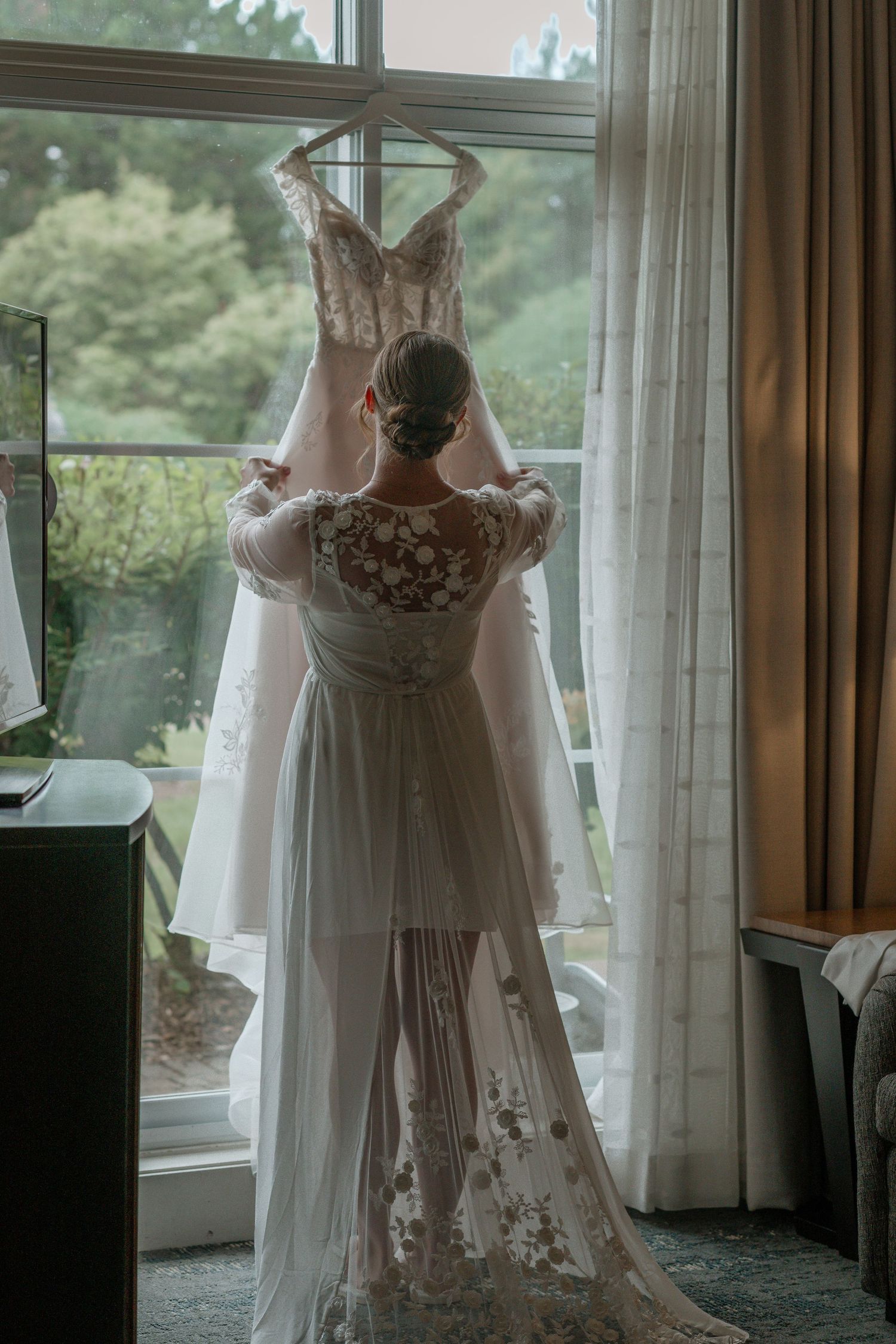A wedding dress hangs in front of a window while someone admires the delicate lace details and flowing train.