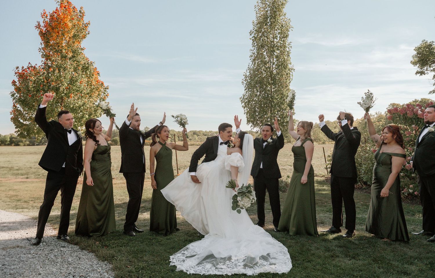 Wedding party poses together on a grassy hillside during golden hour in autumn with colorful trees in background.