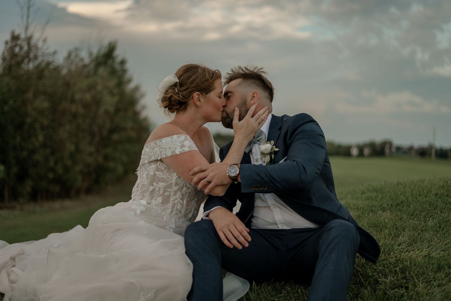 A romantic wedding couple embraces in a lush green field under a dramatic cloudy sky.