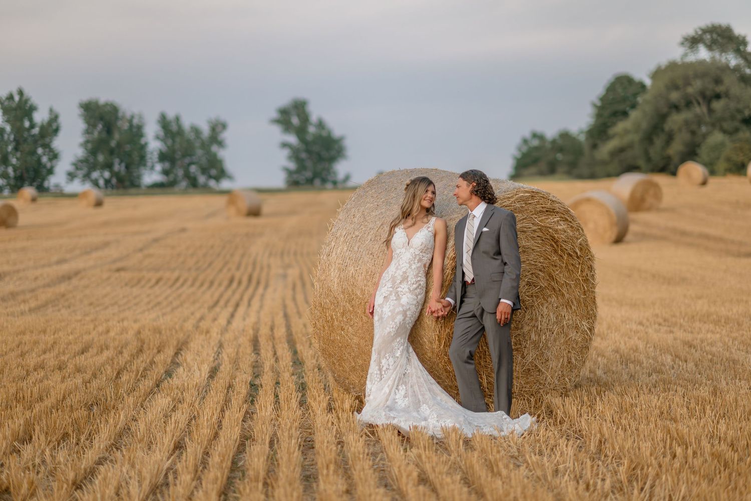 Newlyweds stand beside a round hay bale in a harvested field with trees in the background.