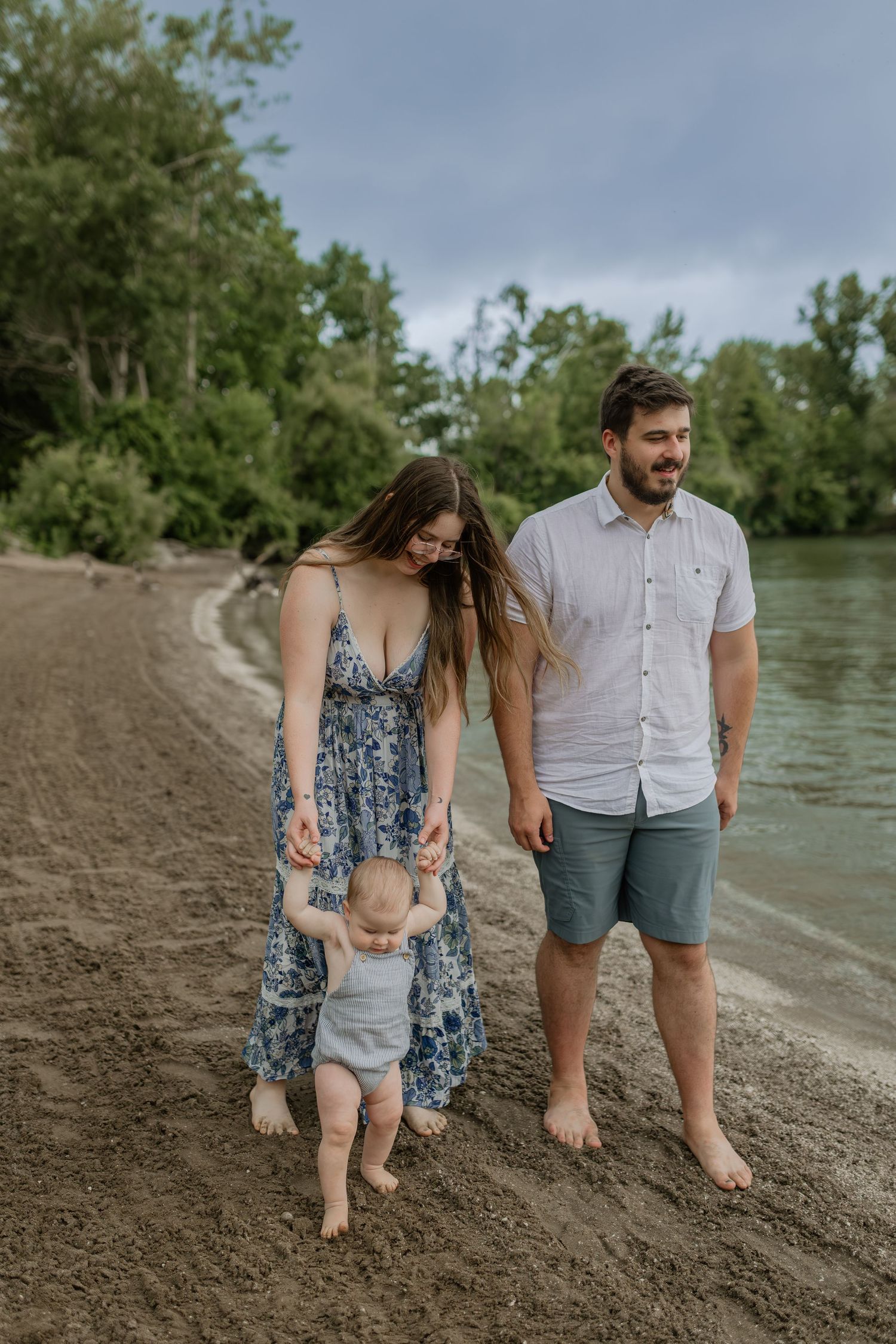 Couple walking barefoot along muddy riverbank on cloudy summer day.