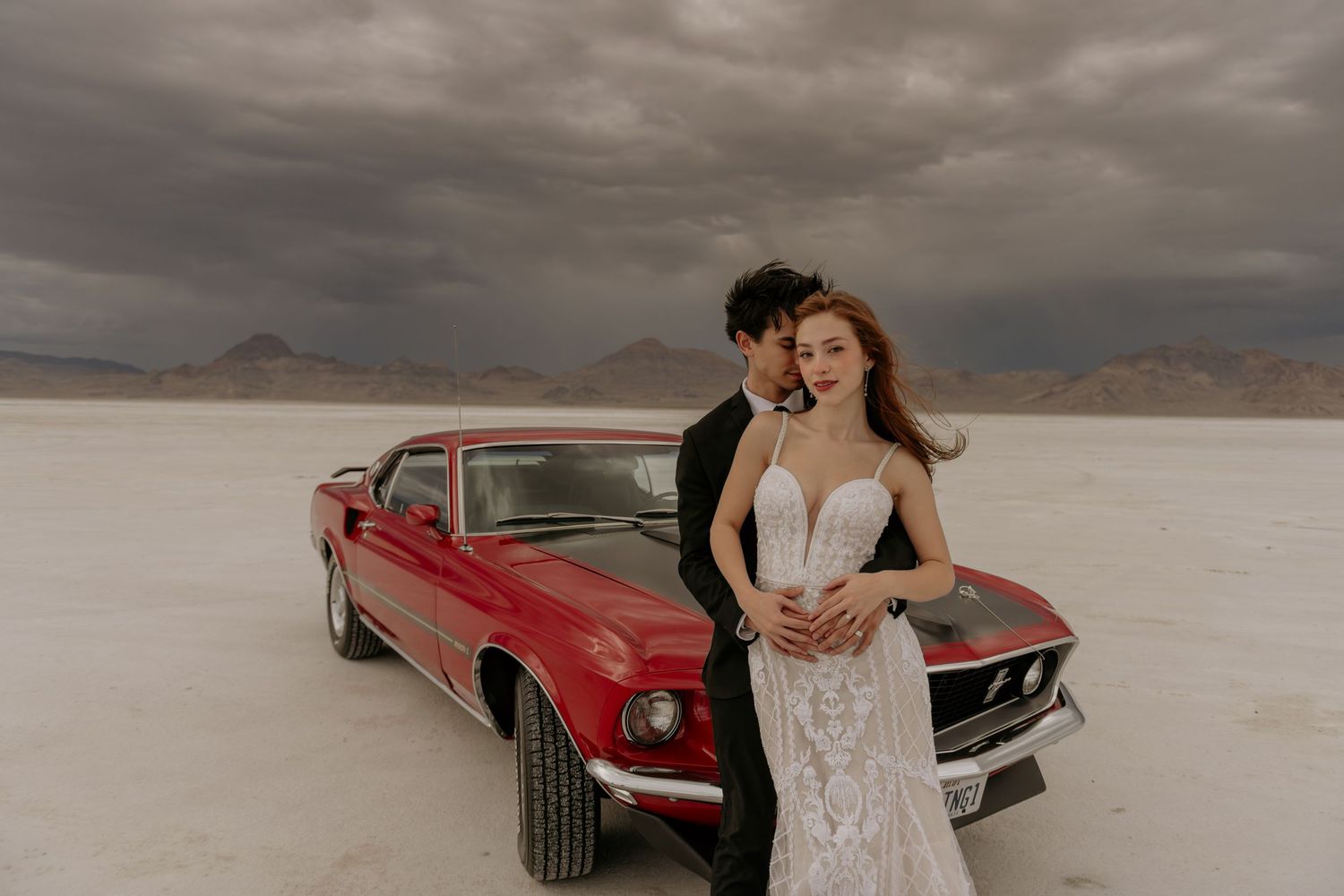 A couple in wedding attire embraces next to a vintage red Ford Mustang on a salt flat under dramatic stormy skies.