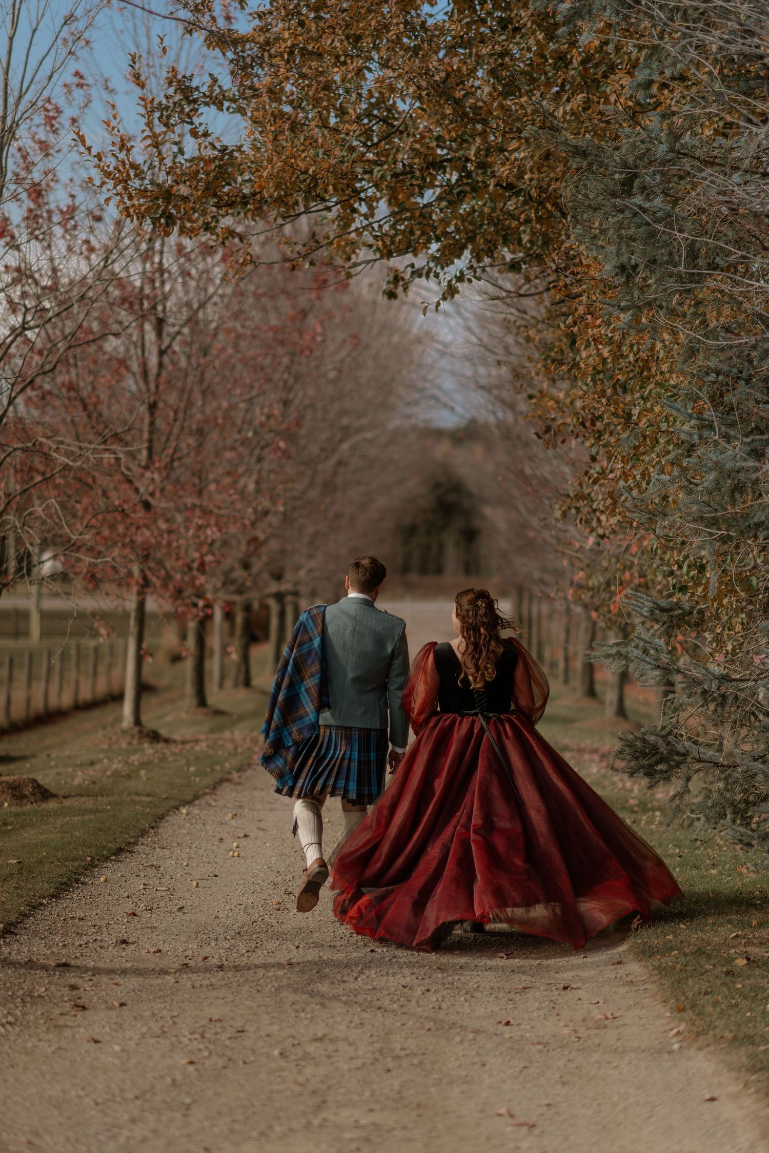 Couple in vintage formal attire walk down tree-lined autumn path with burgundy dress flowing in motion.