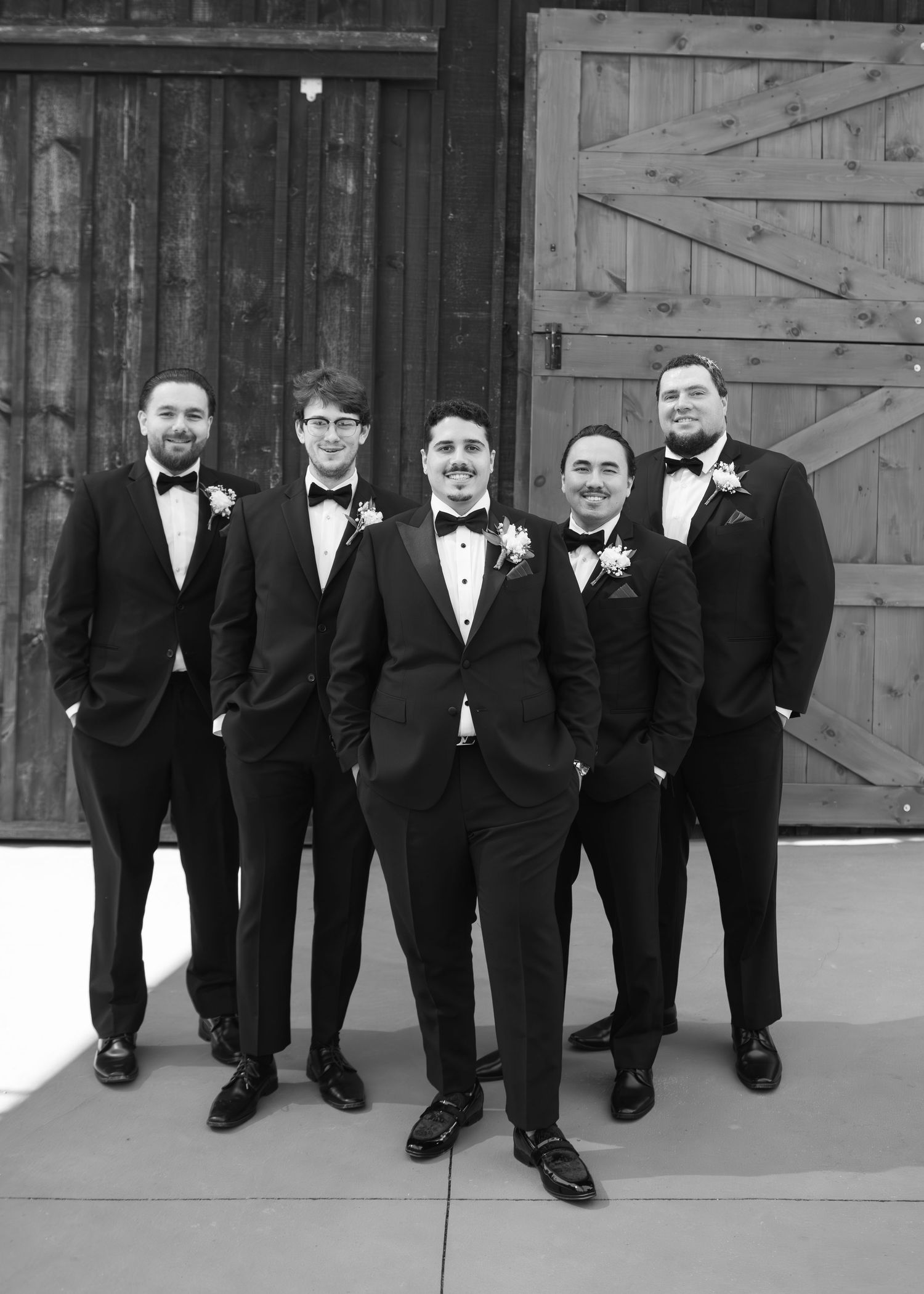 Black and white photo showing groomsmen's legs and dress shoes lined up against a rustic barn wall.