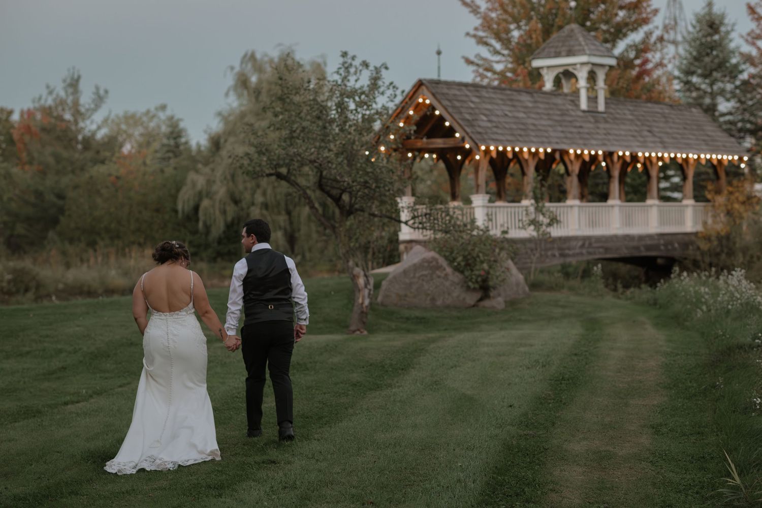 A couple walks down a grassy path at sunset with string lights glowing in the background at an outdoor wedding venue.