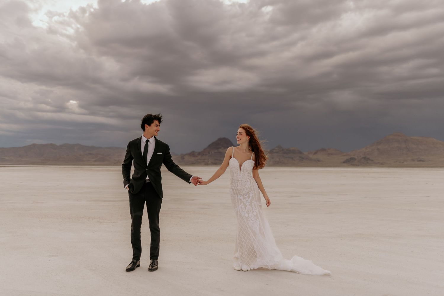 A couple in wedding attire holds hands while standing on a desert landscape under dramatic cloudy skies.