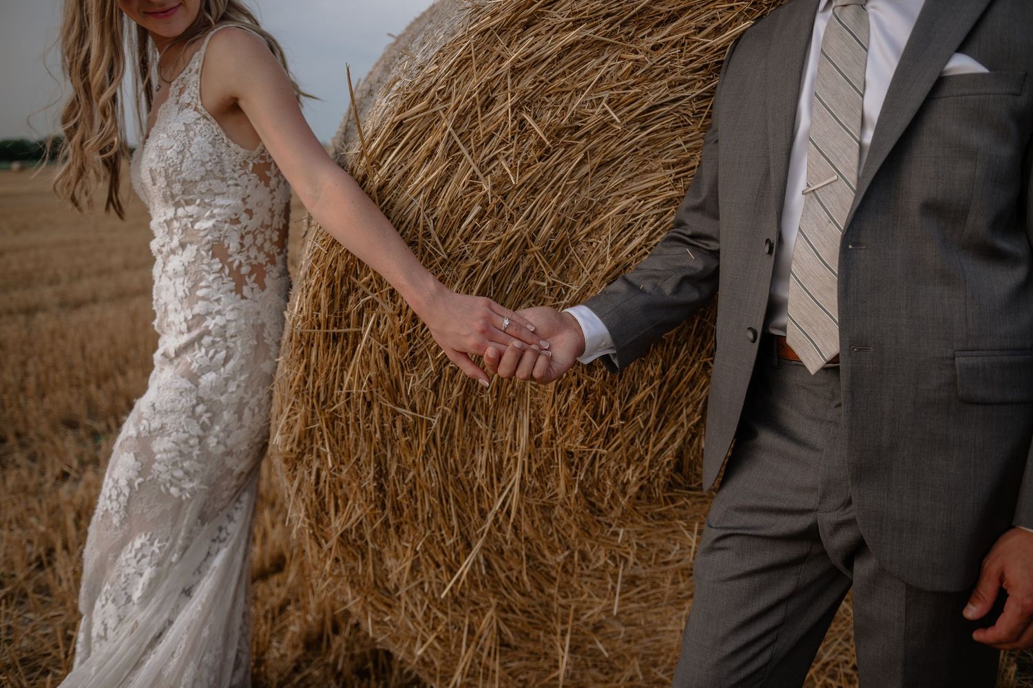 Close-up of joined hands between a bride in lace and groom in grey suit against hay bale backdrop.