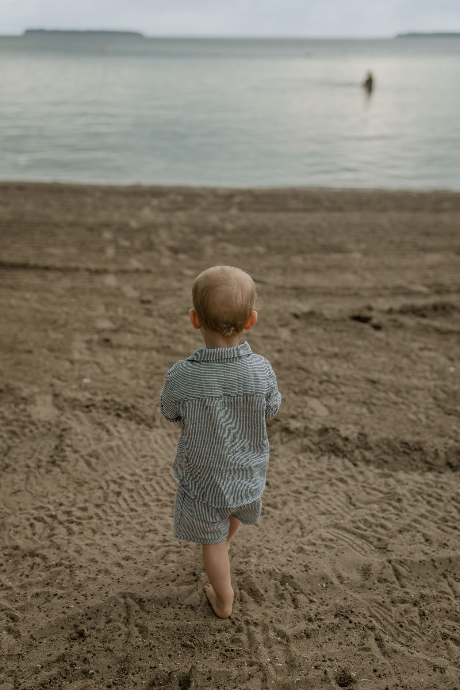 Small child in blue shirt walking alone on a sandy beach with ocean waves in the background.