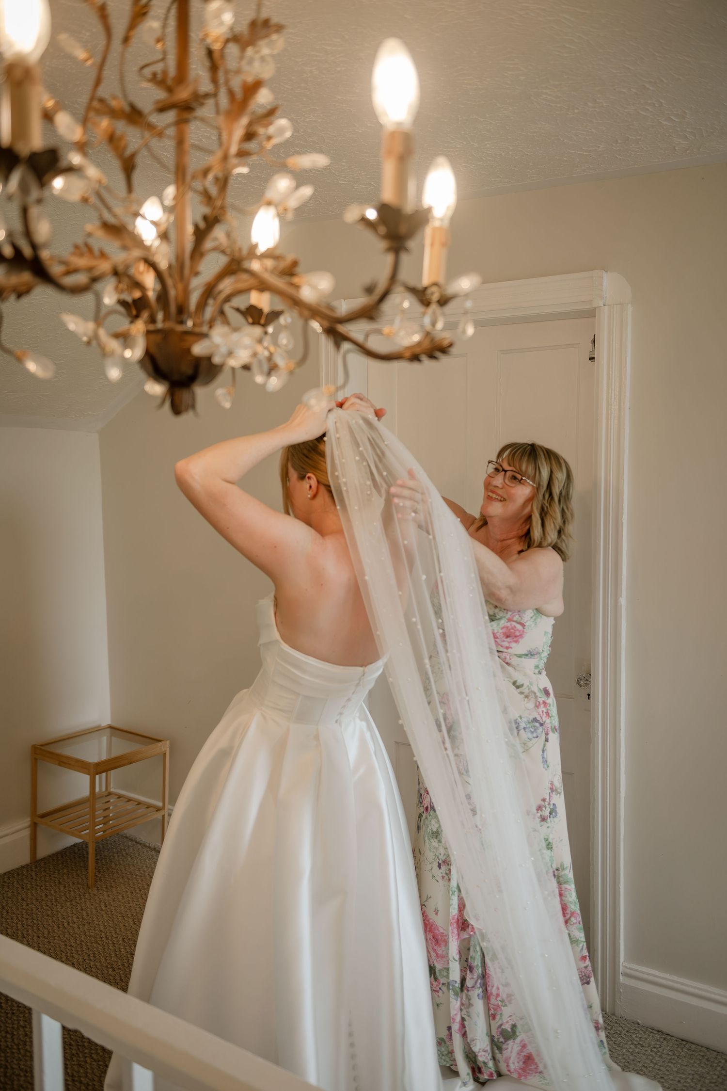 A wedding dress hangs from an ornate chandelier in a bright white room while being prepared for a special day. Mom, puts on a veil