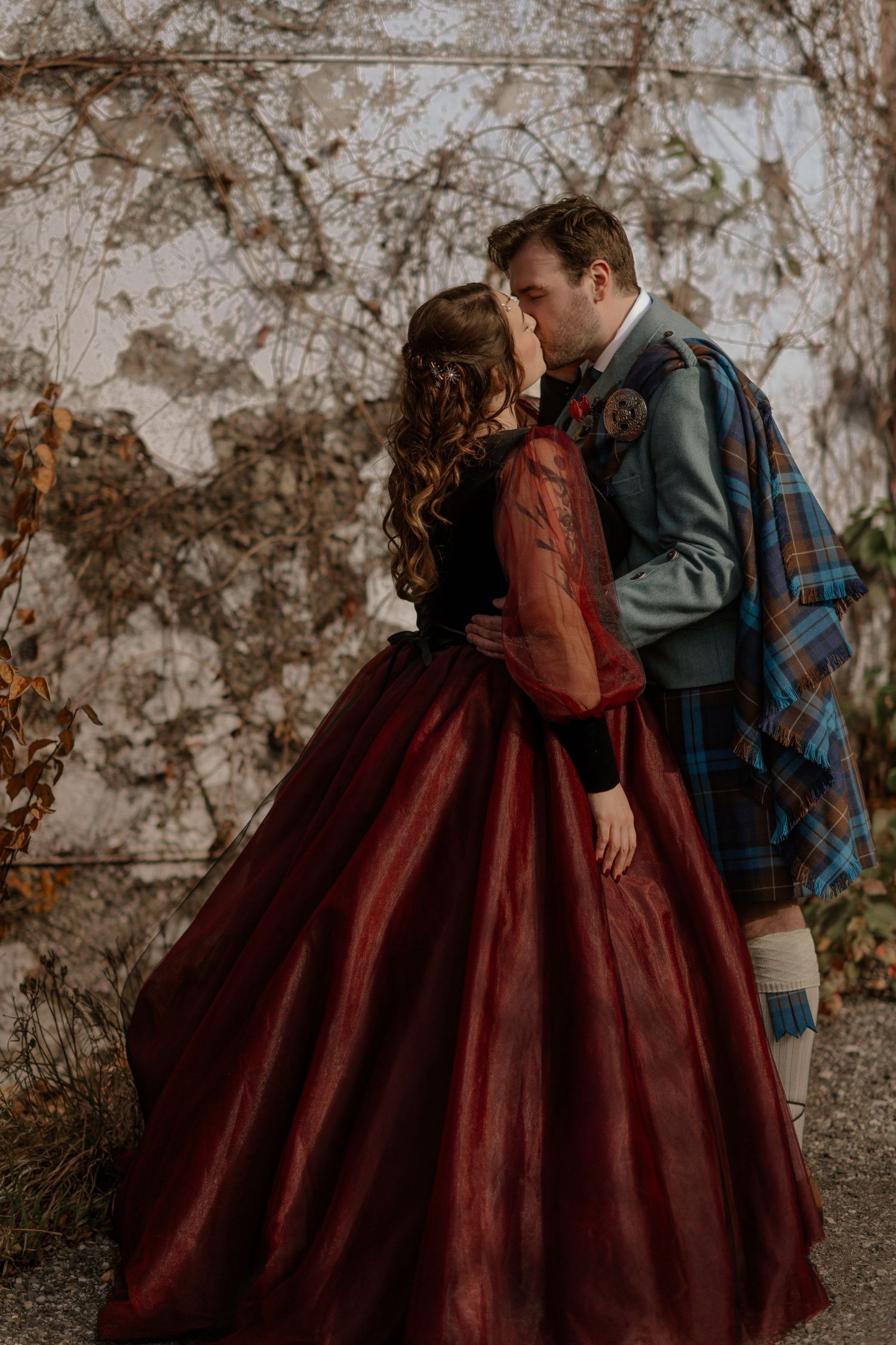 A romantic couple in Scottish attire share an intimate moment against a textured wall in autumn.