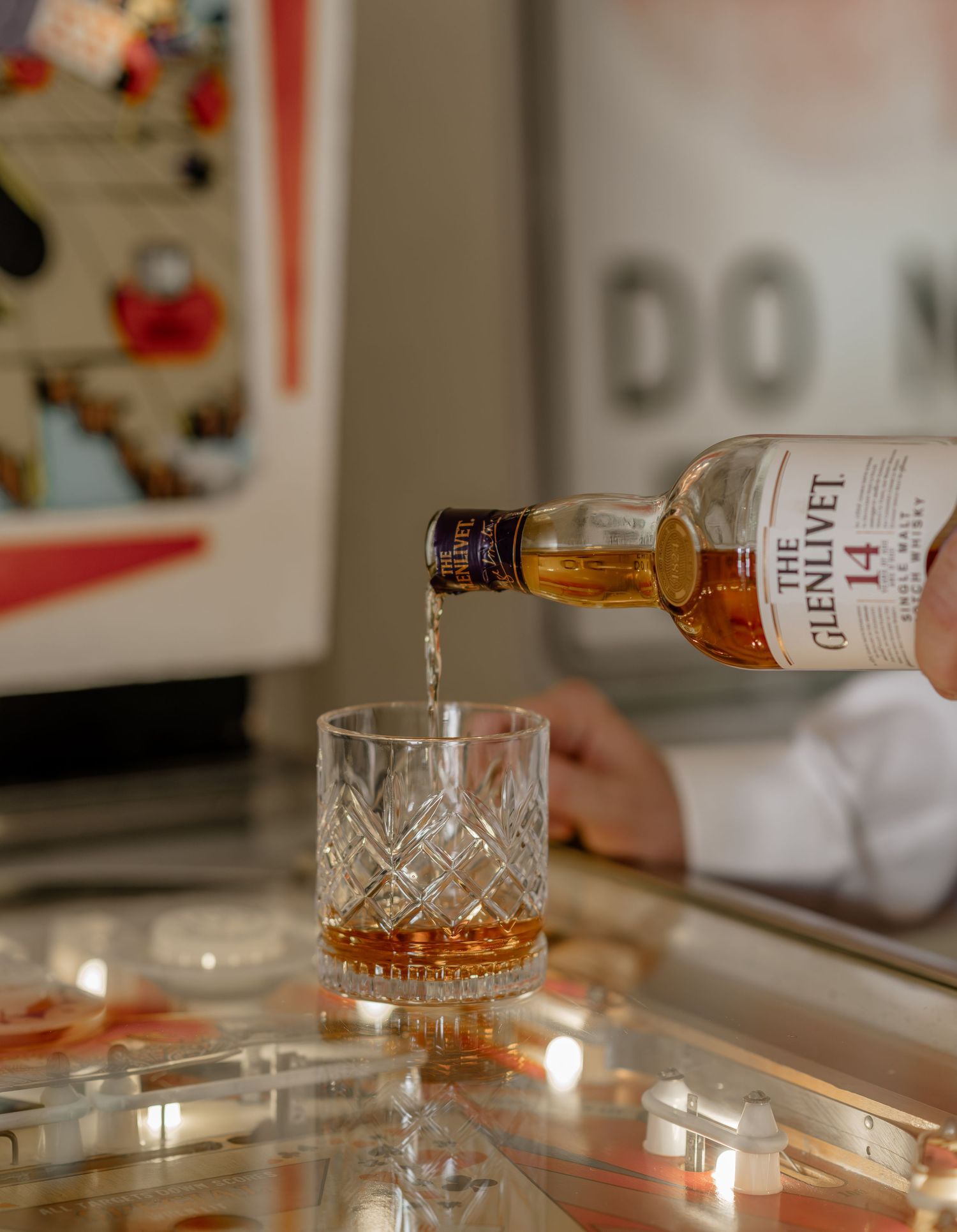 Crystal glasses being filled with amber whiskey on a white countertop in warm lighting.