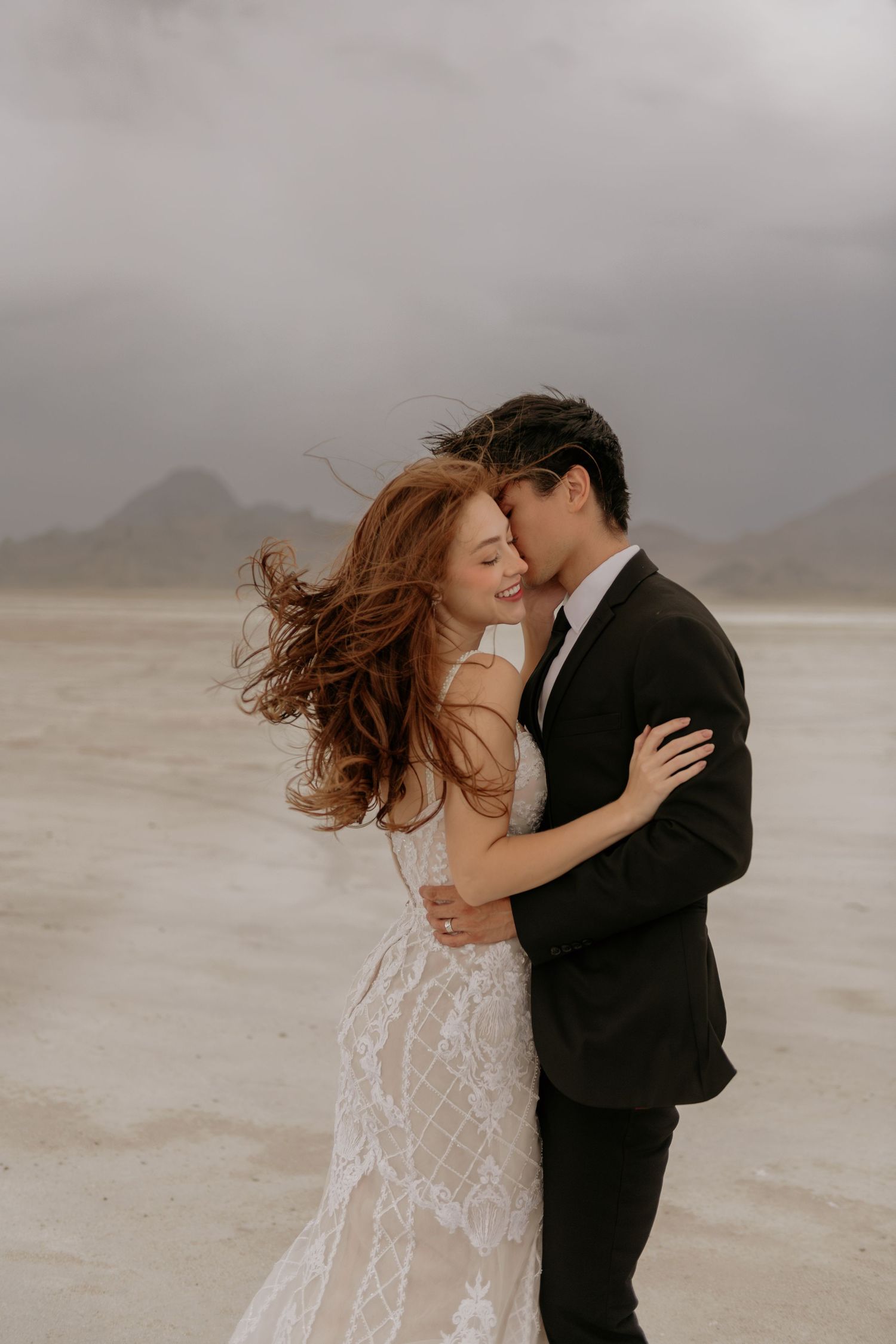 A romantic couple embraces and kisses on a misty beach while wearing formal wedding attire.
