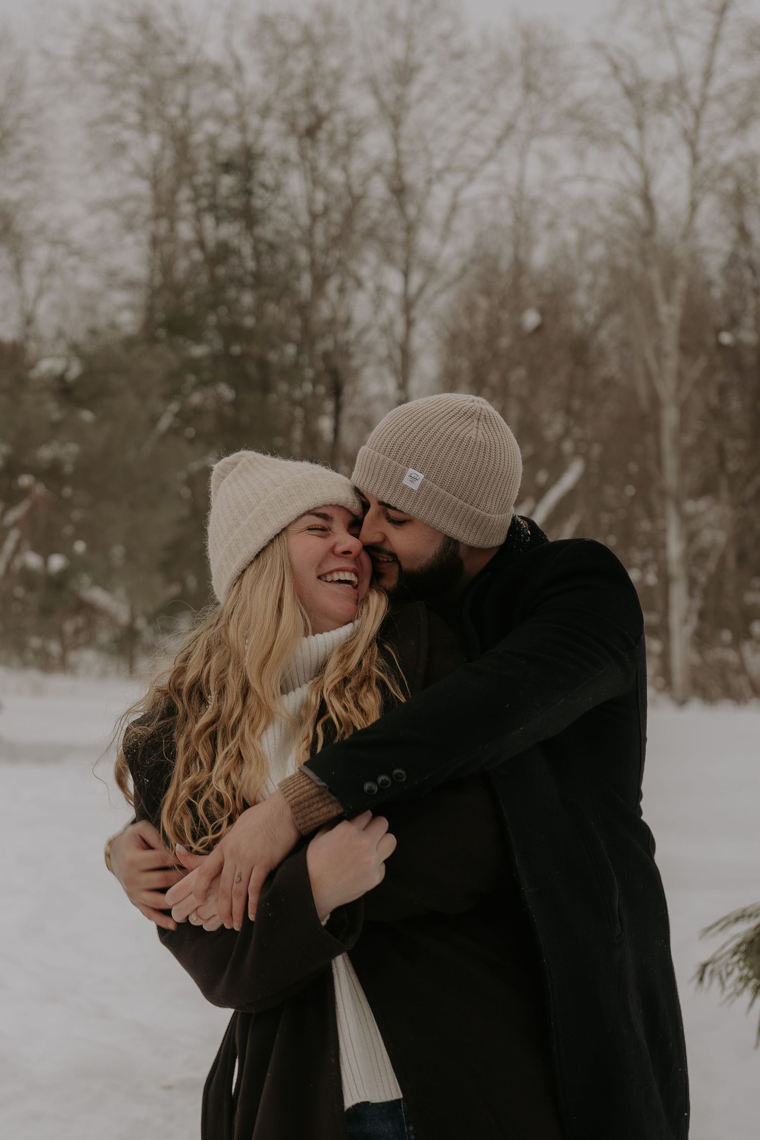 Couple laughing and embracing in a snowy forest while wearing winter clothing.
