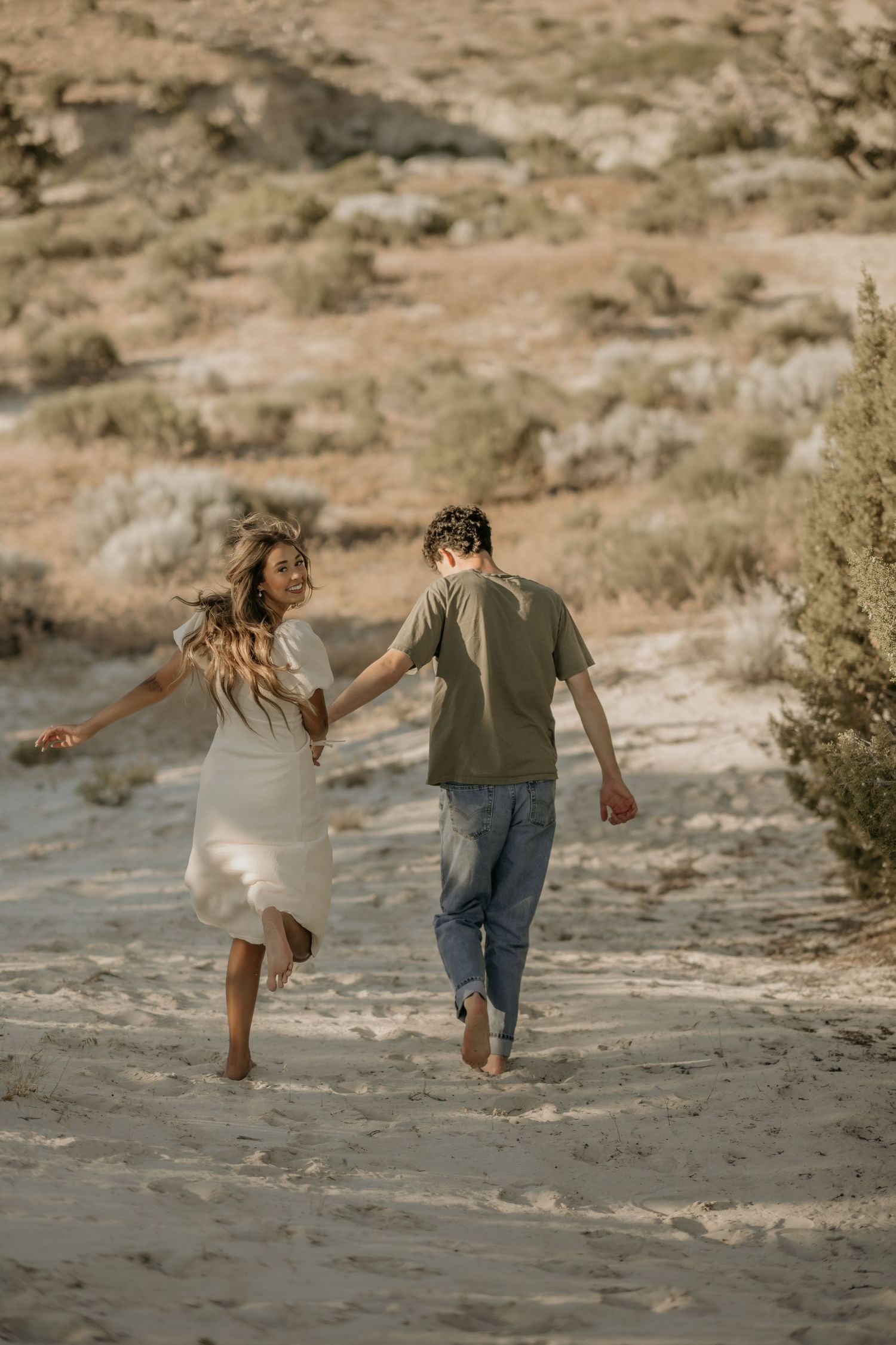 A couple runs playfully along a sandy desert landscape during golden hour.