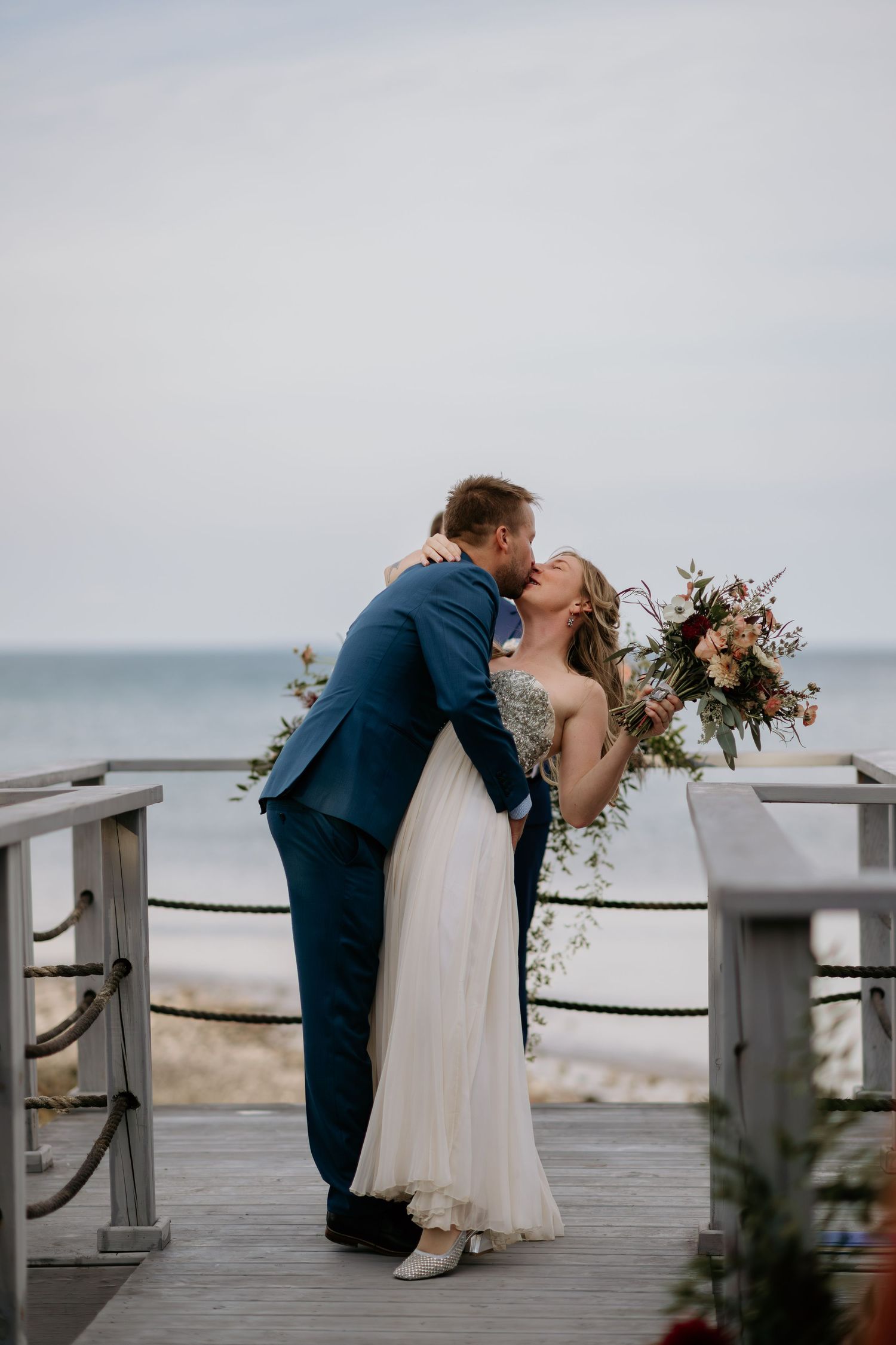 A romantic wedding couple shares a passionate kiss on a coastal boardwalk overlooking the ocean at dusk.