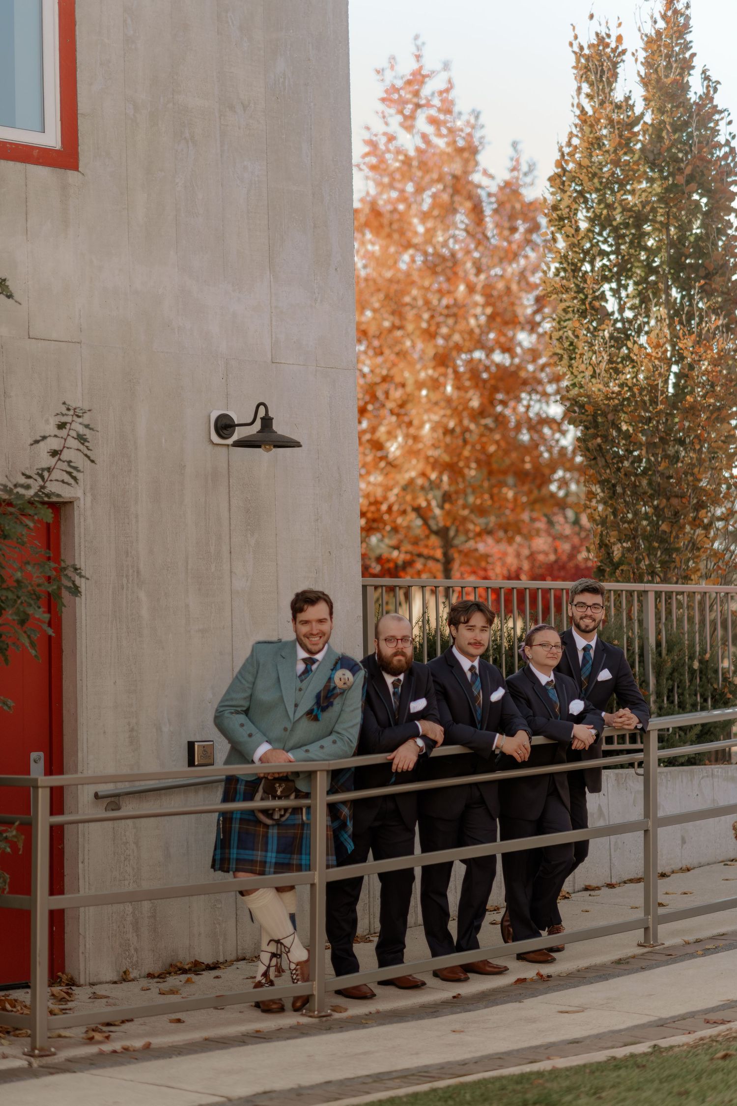A group of people lean against a railing outside a building on an autumn day with orange trees in the background.