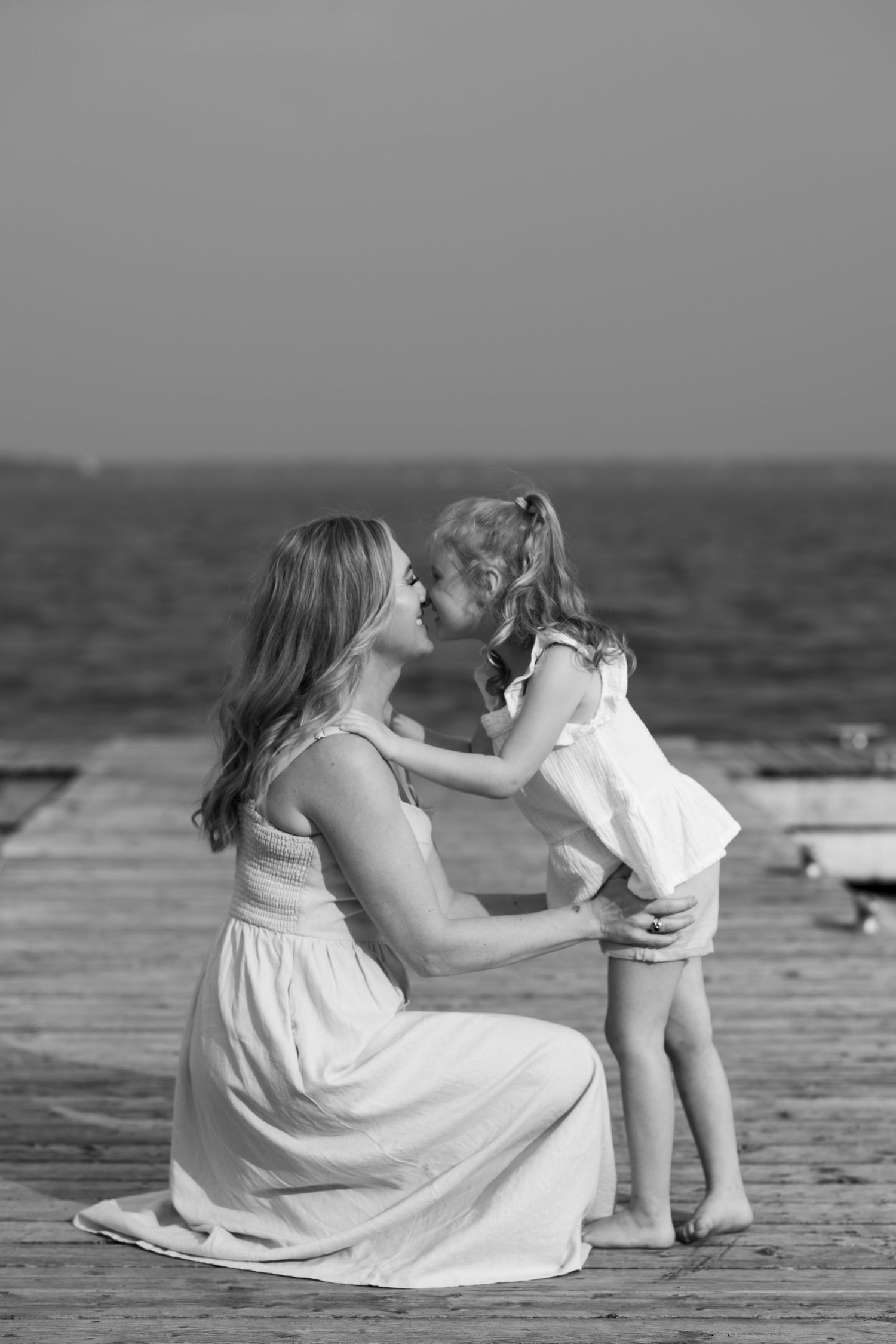 A tender moment between parent and child sitting by the ocean in white summer clothing.