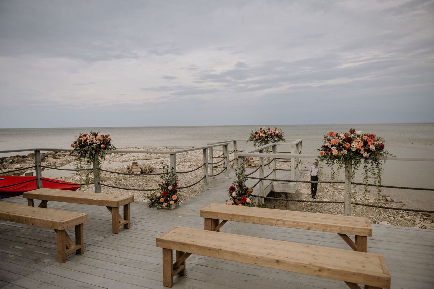 Rustic wooden benches and floral arrangements line a beachfront ceremony setup with rope barriers and ocean views.