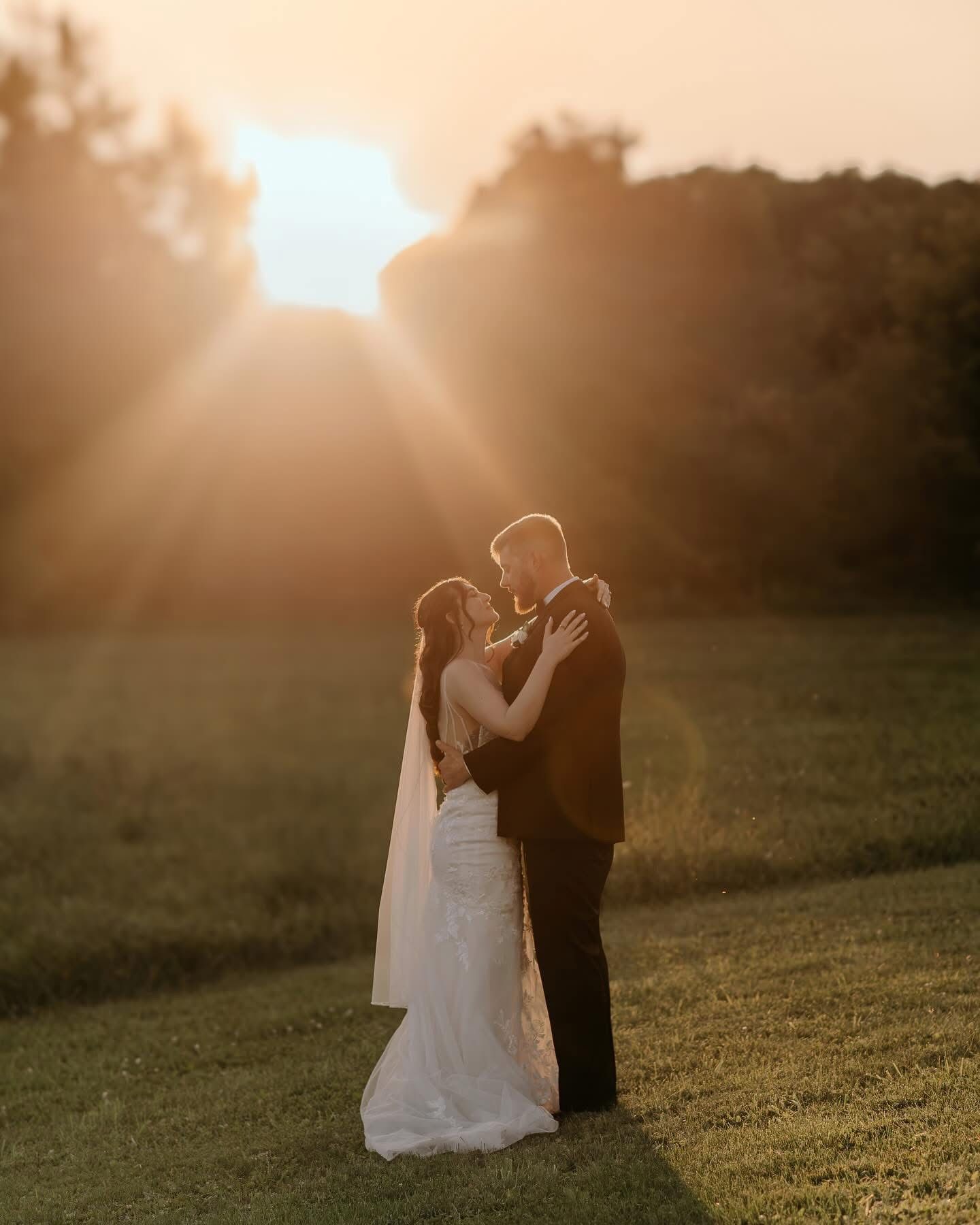 A couple shares an intimate moment at sunset in a grassy field, with golden sunbeams streaming through trees.