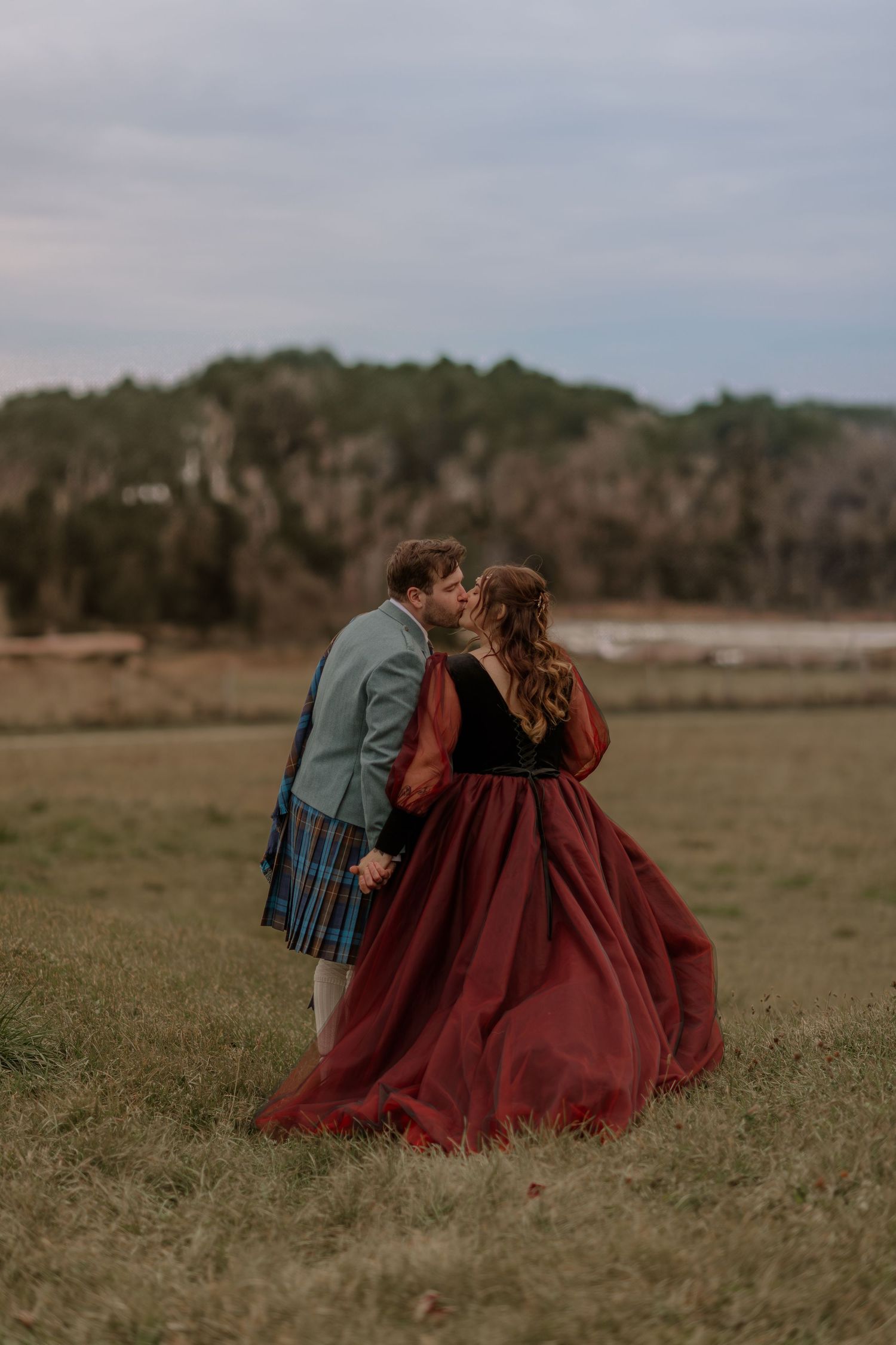 A couple in burgundy dress and blue suit sits together in a sunlit meadow.