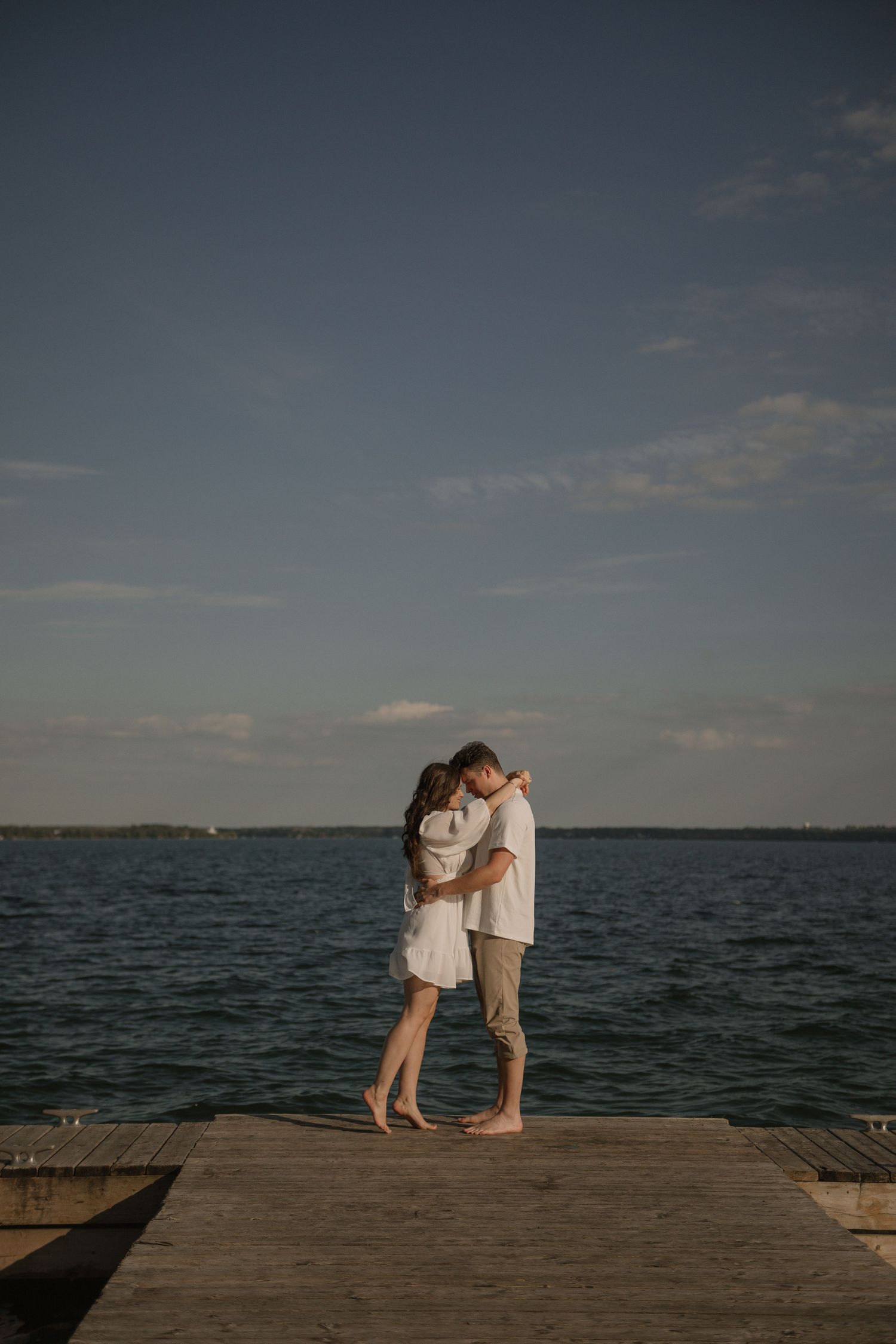 A couple shares an intimate moment on a dock against a moody waterfront backdrop.