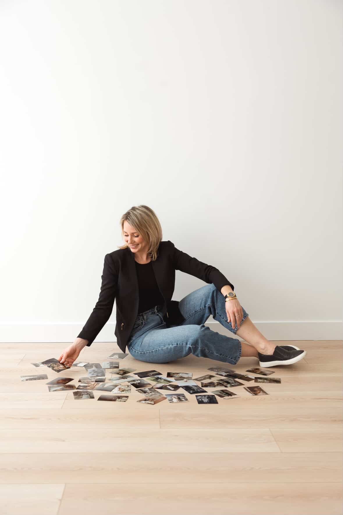 A person in black attire lies on a wooden floor looking at scattered photographs spread around them.