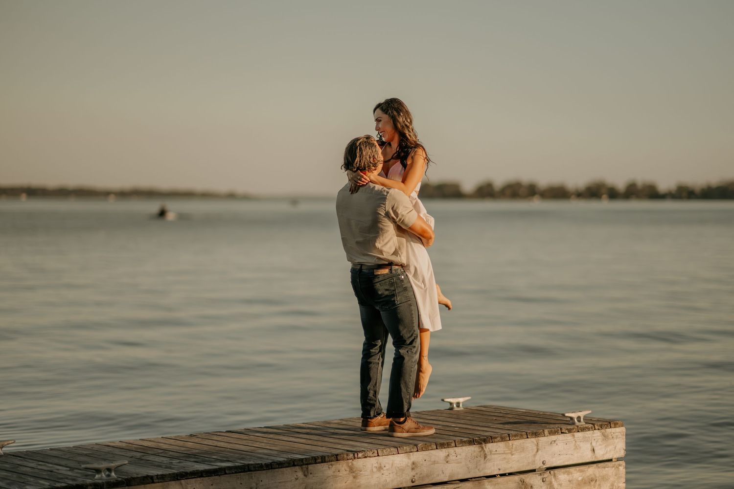 A couple embraces romantically on a wooden dock overlooking a serene lake at sunset.