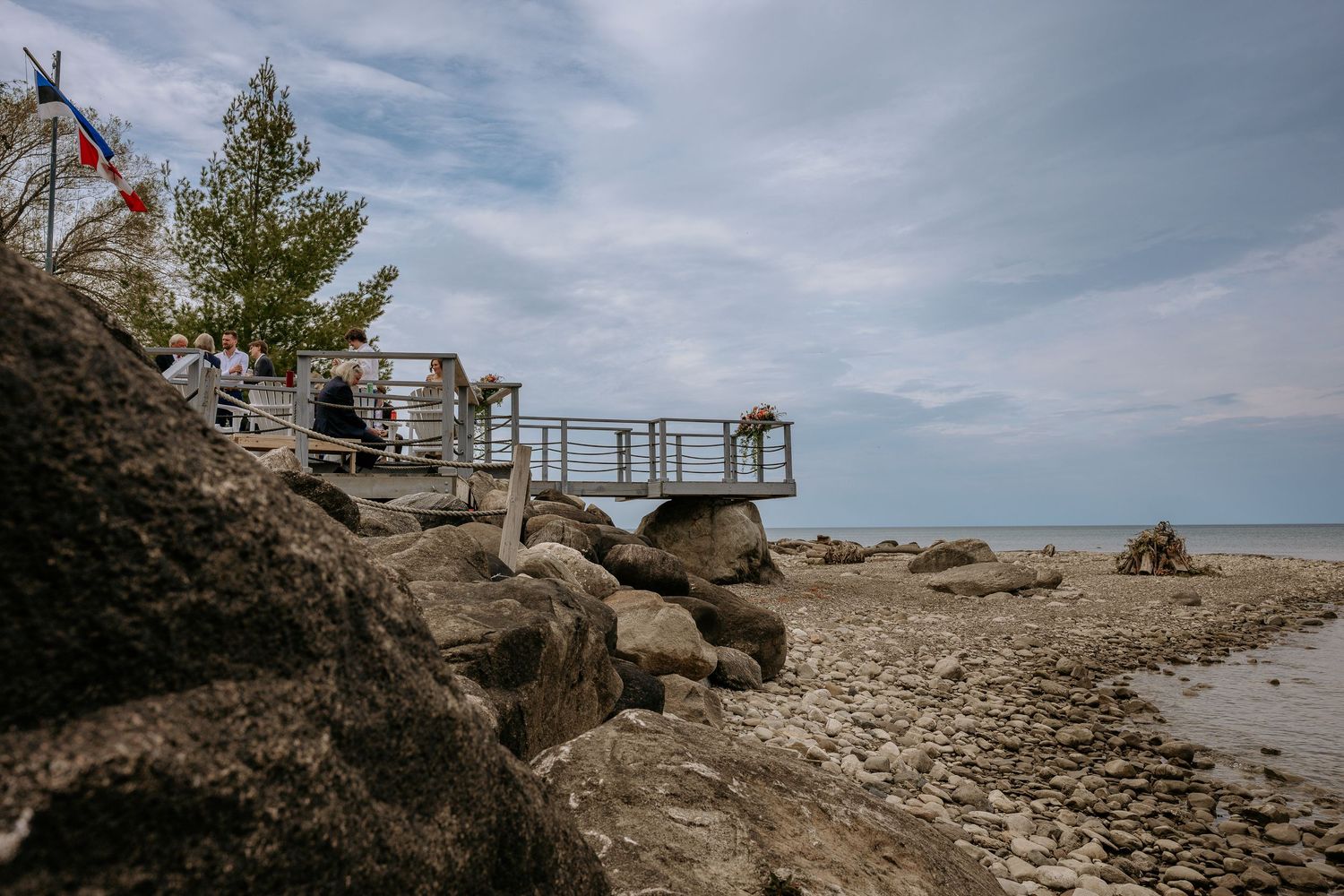 A wooden viewing platform built on rocky coastal terrain overlooks a beach under cloudy skies.