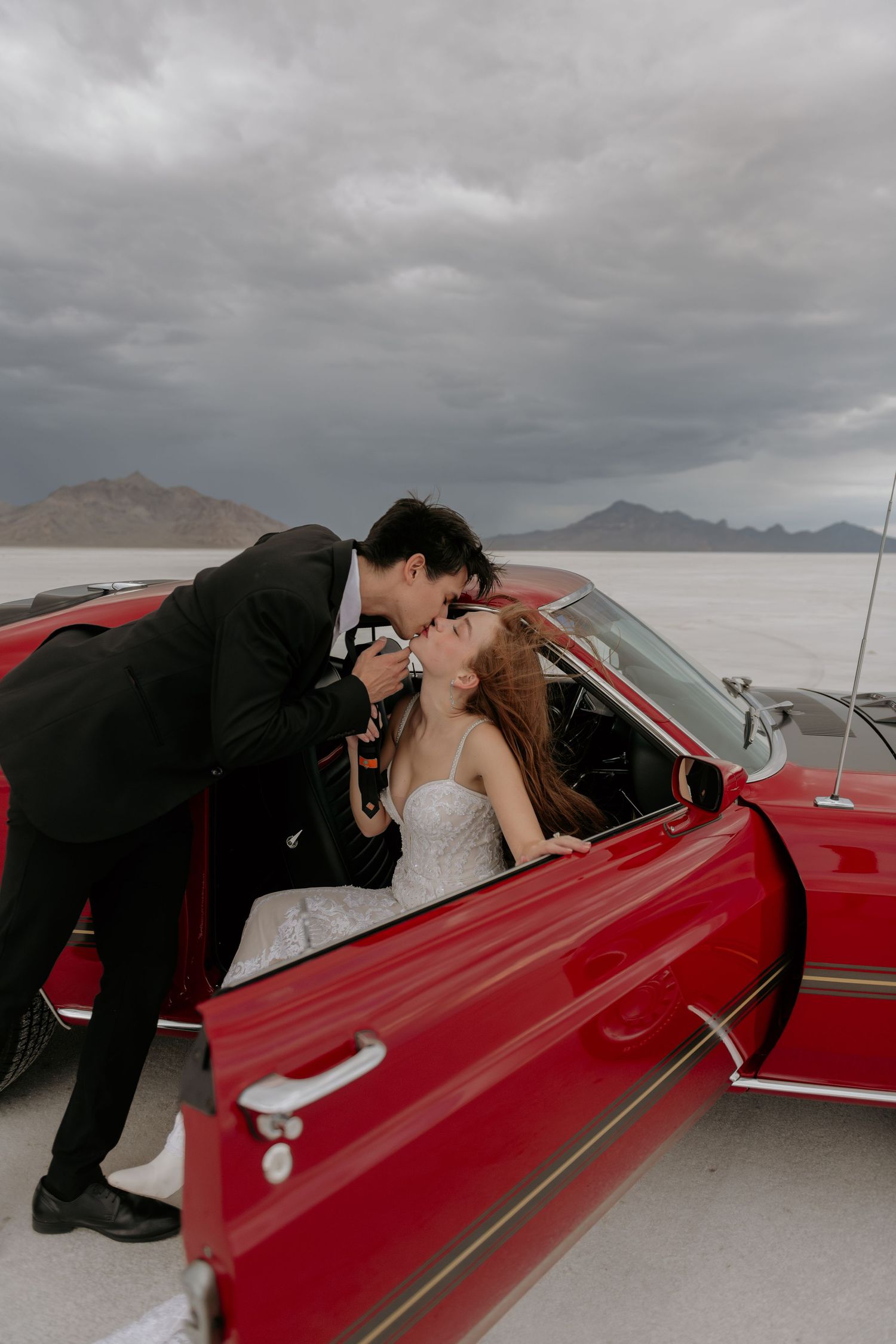 A couple shares a romantic kiss in a vintage red car against a dramatic cloudy desert landscape at Bonneville Salt Flats.