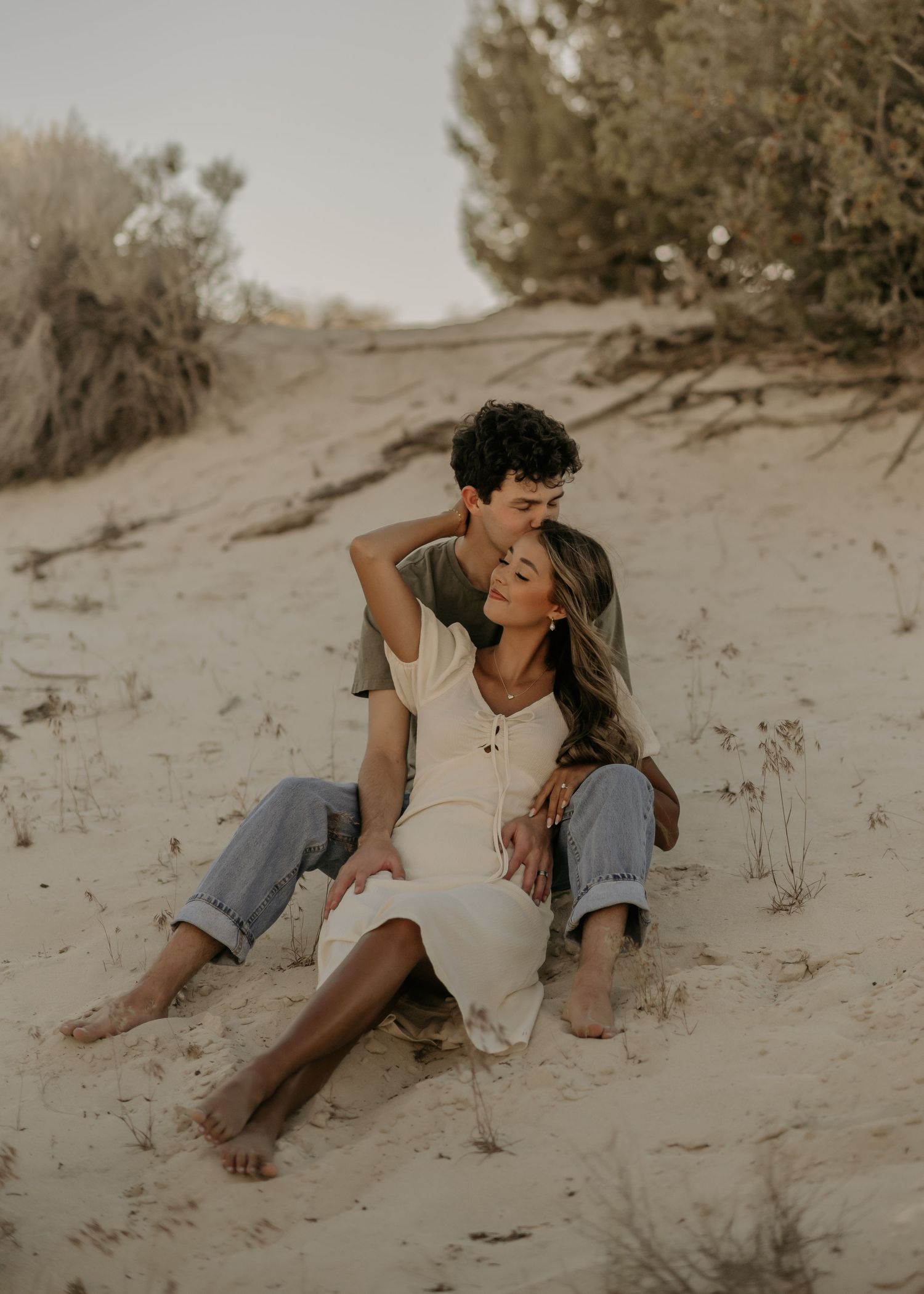 A couple sits together on sandy dunes, sharing intimate moments in a desert landscape during golden hour.