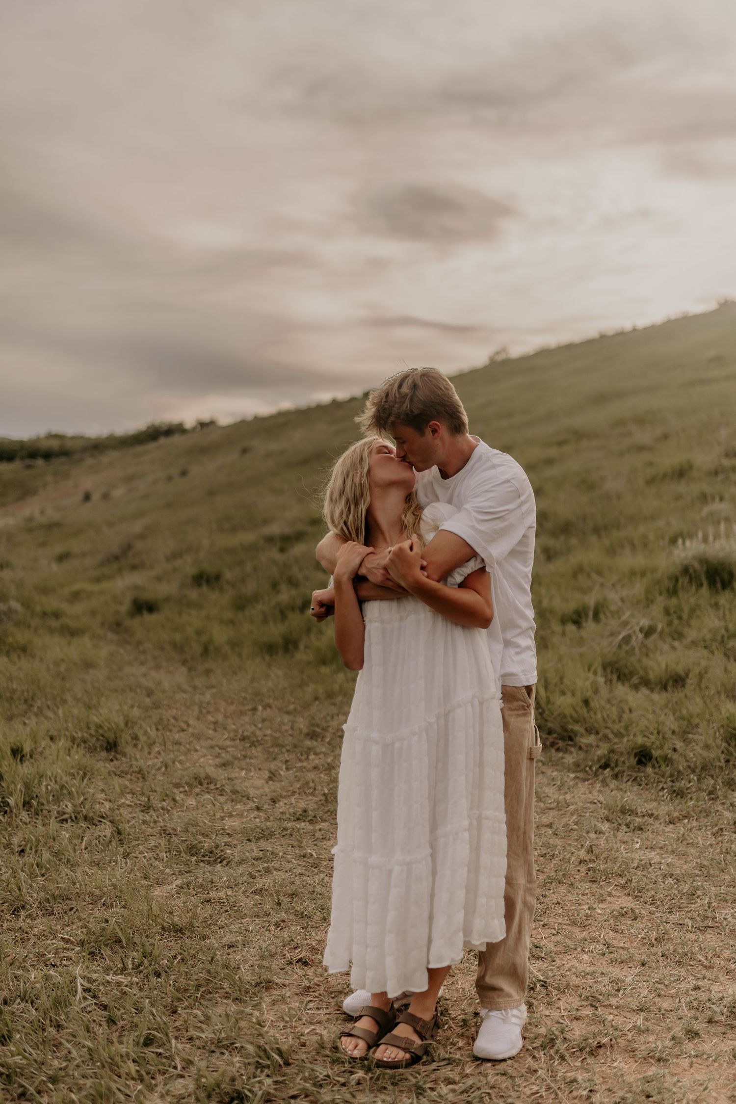 A couple shares tender moments on a grassy hillside during a romantic outdoor photoshoot at sunset.