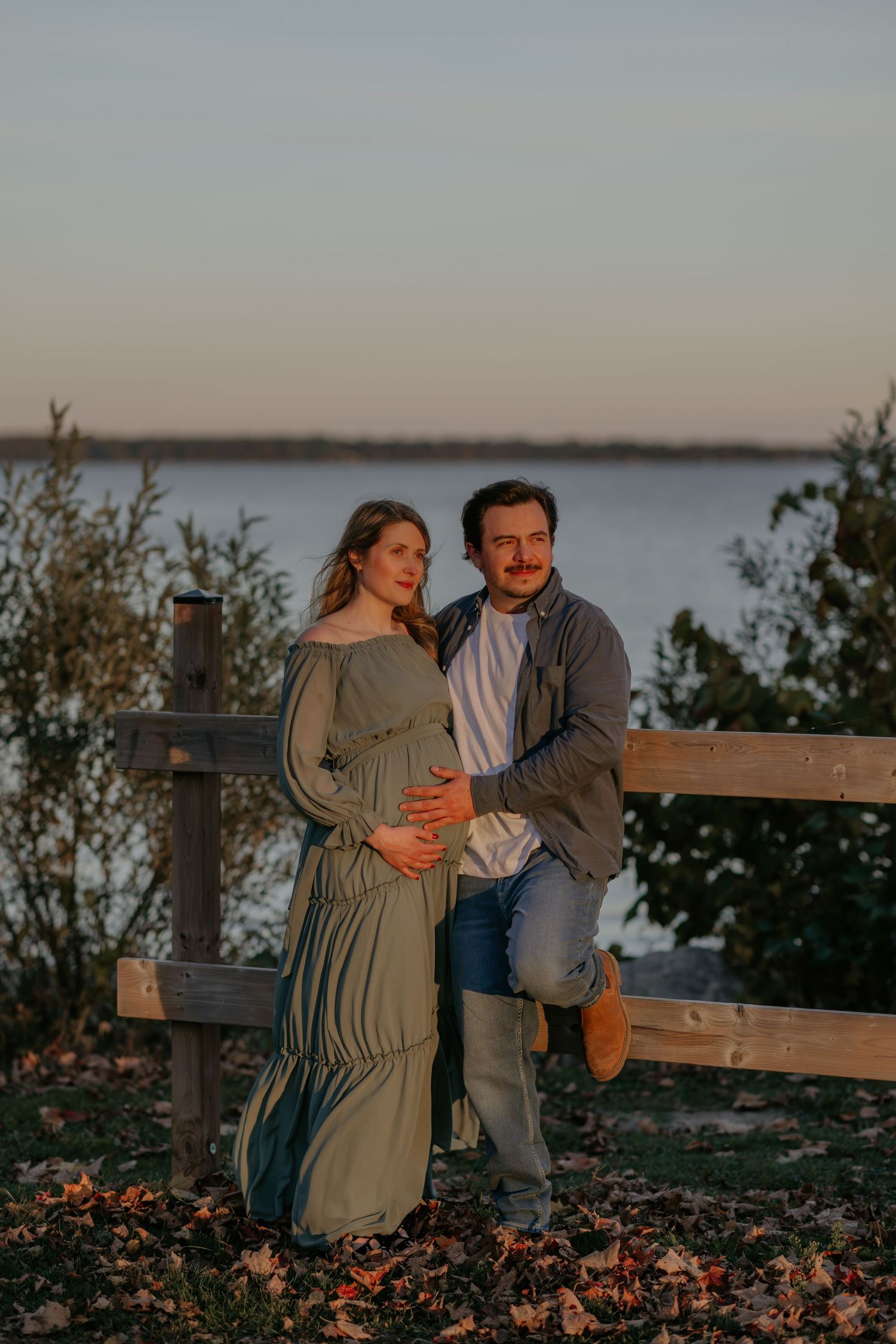 Sunset maternity photos by waterfront with couple on wooden bench surrounded by fall foliage.